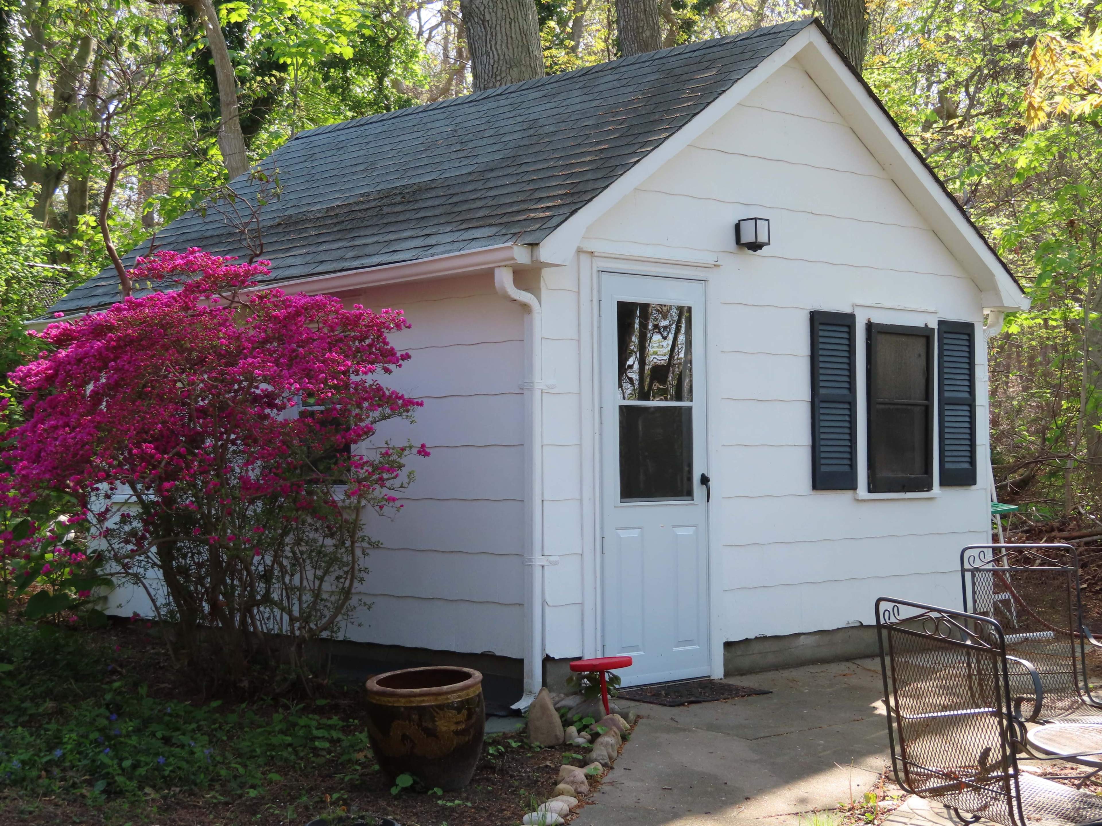 A small white cottage with a slanted roof is surrounded by greenery and features a vibrant flowering bush in front.