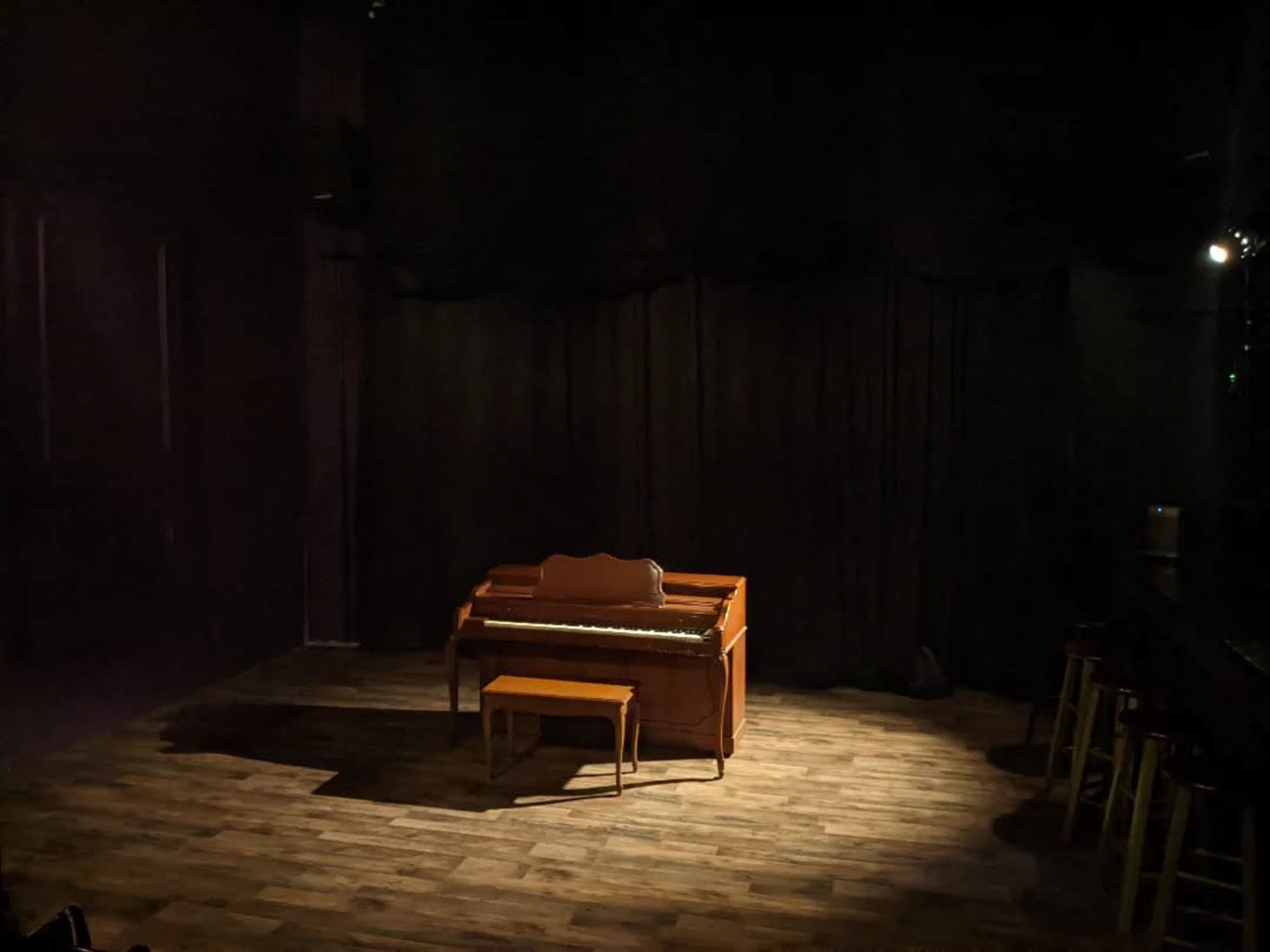 A dimly lit stage with a wooden piano and a small chair set against a backdrop of black curtains.