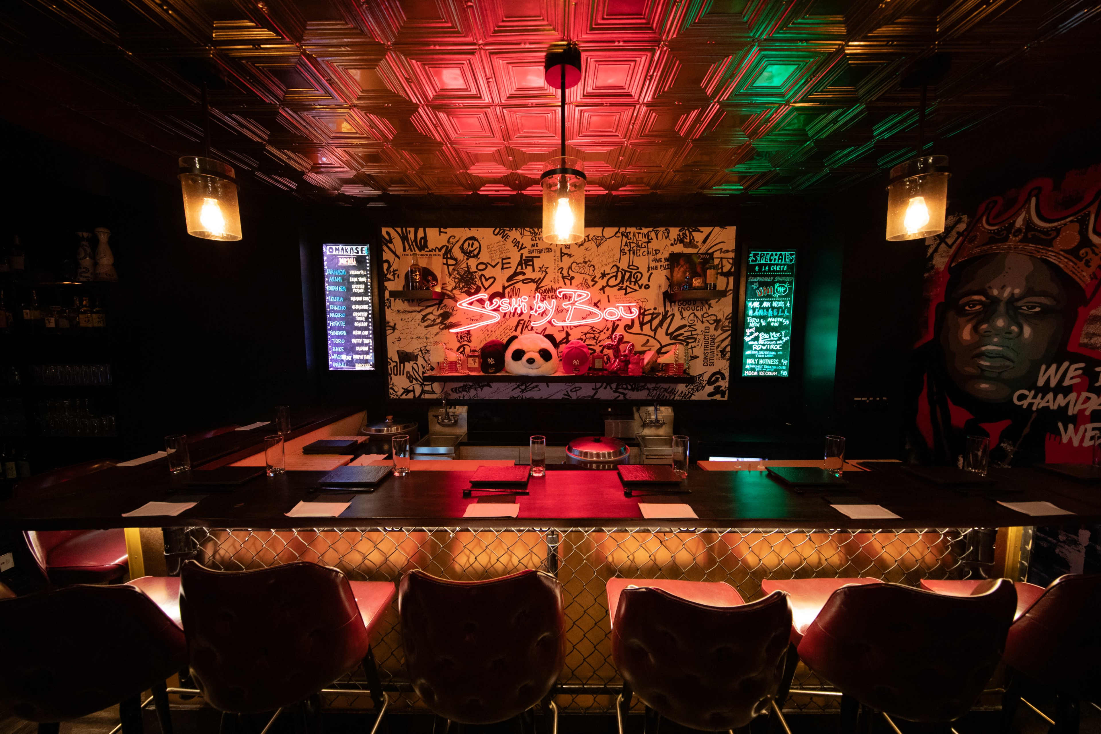 The image shows a dimly lit bar with a stylish counter, graffiti art on the wall, and plush seating.