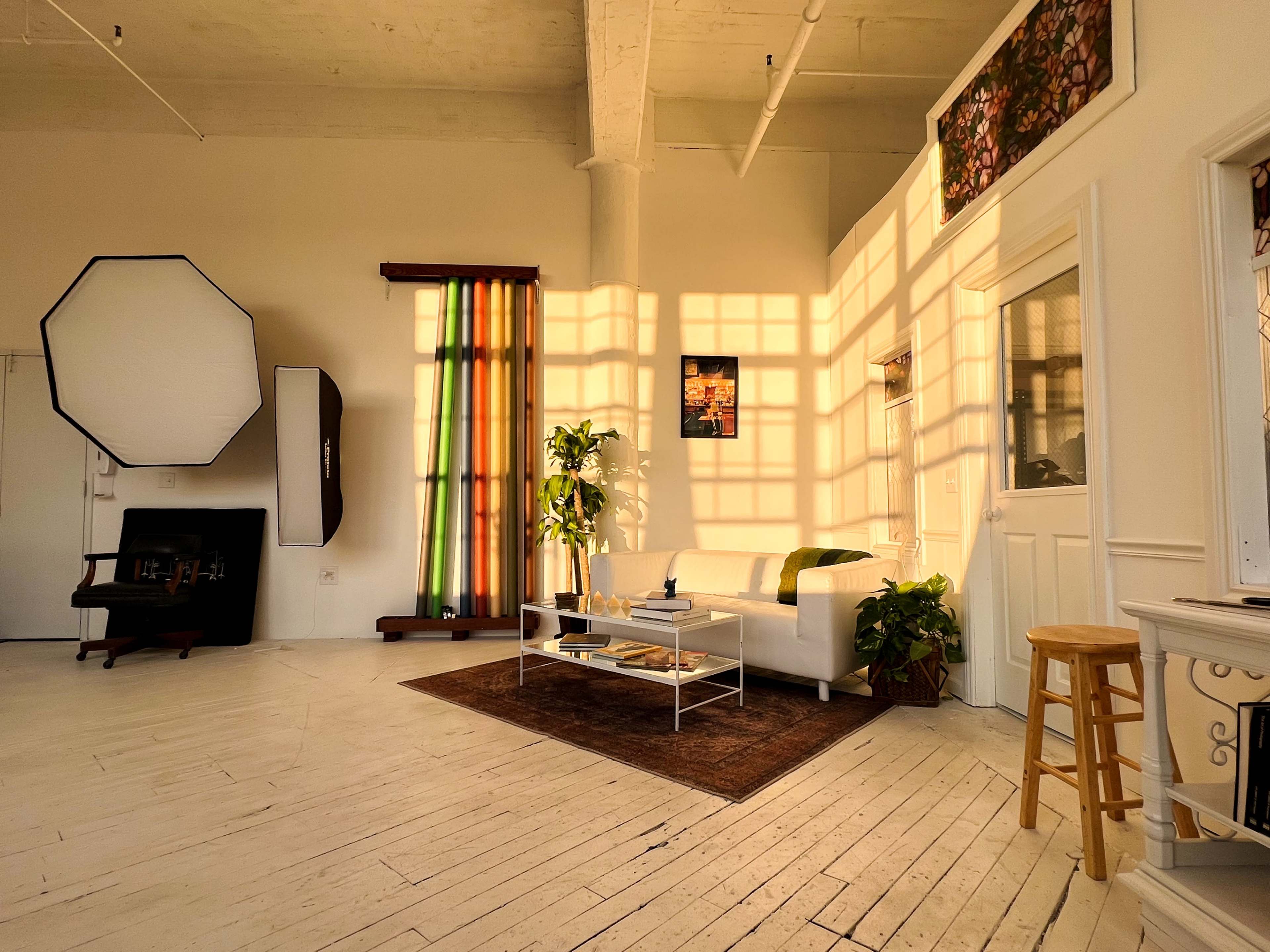 The image shows a bright, modern living space with a white sofa, a coffee table stacked with books, and large windows casting patterned shadows on the floor.