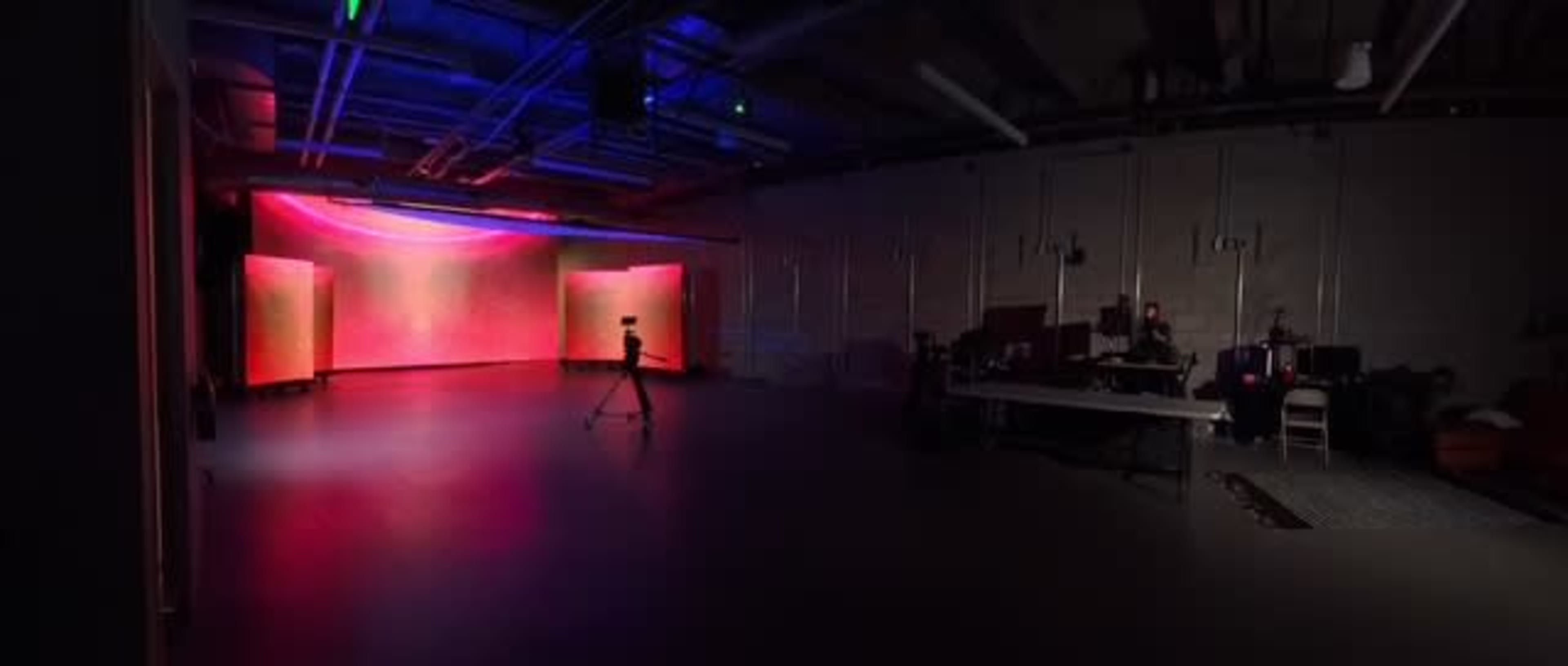 A dimly lit studio features colorful background lights, a camera on a tripod, and equipment setups along one wall.