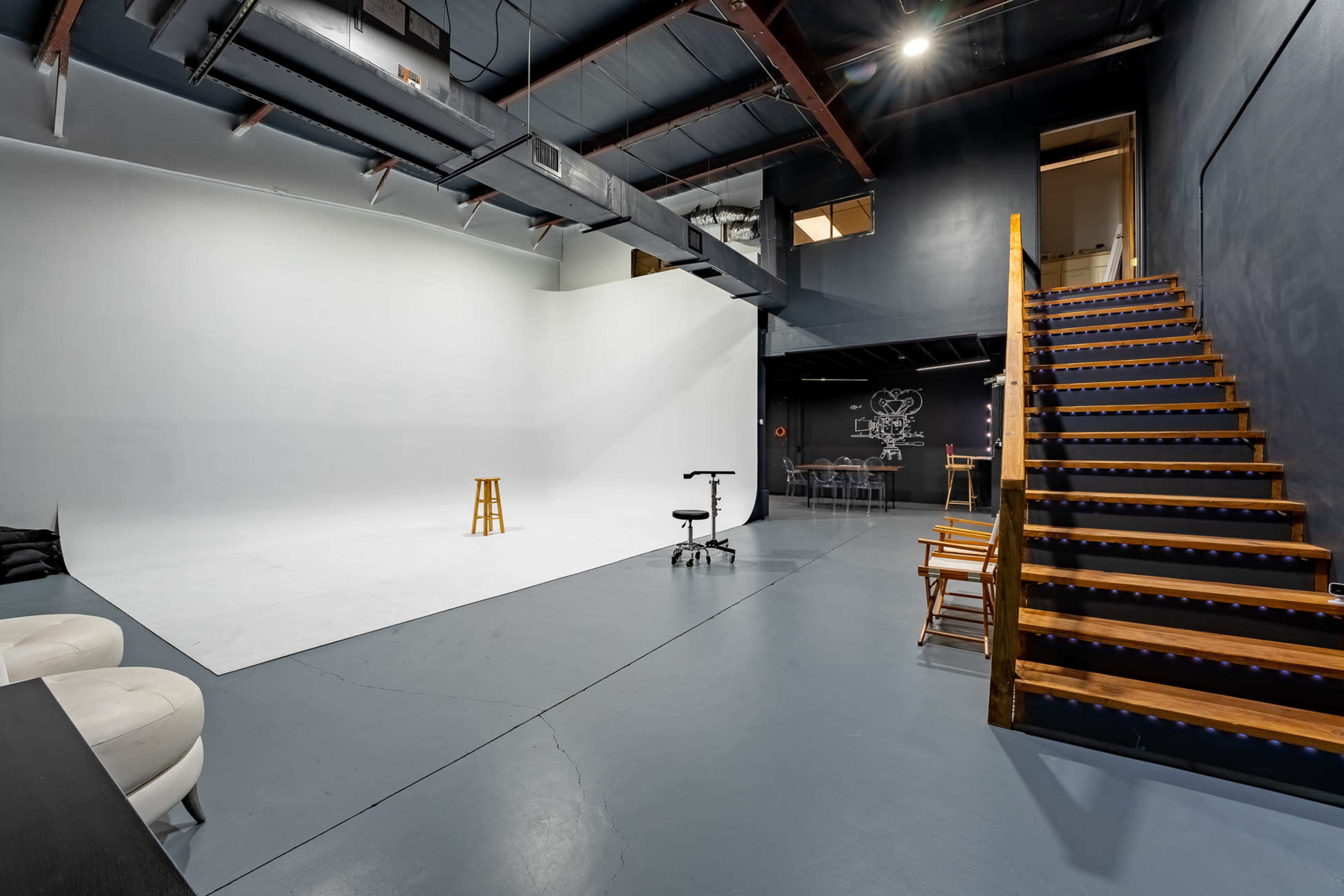 A spacious studio with a white backdrop, wooden stairs leading to an upper level, and various furniture arranged for shooting or meetings.