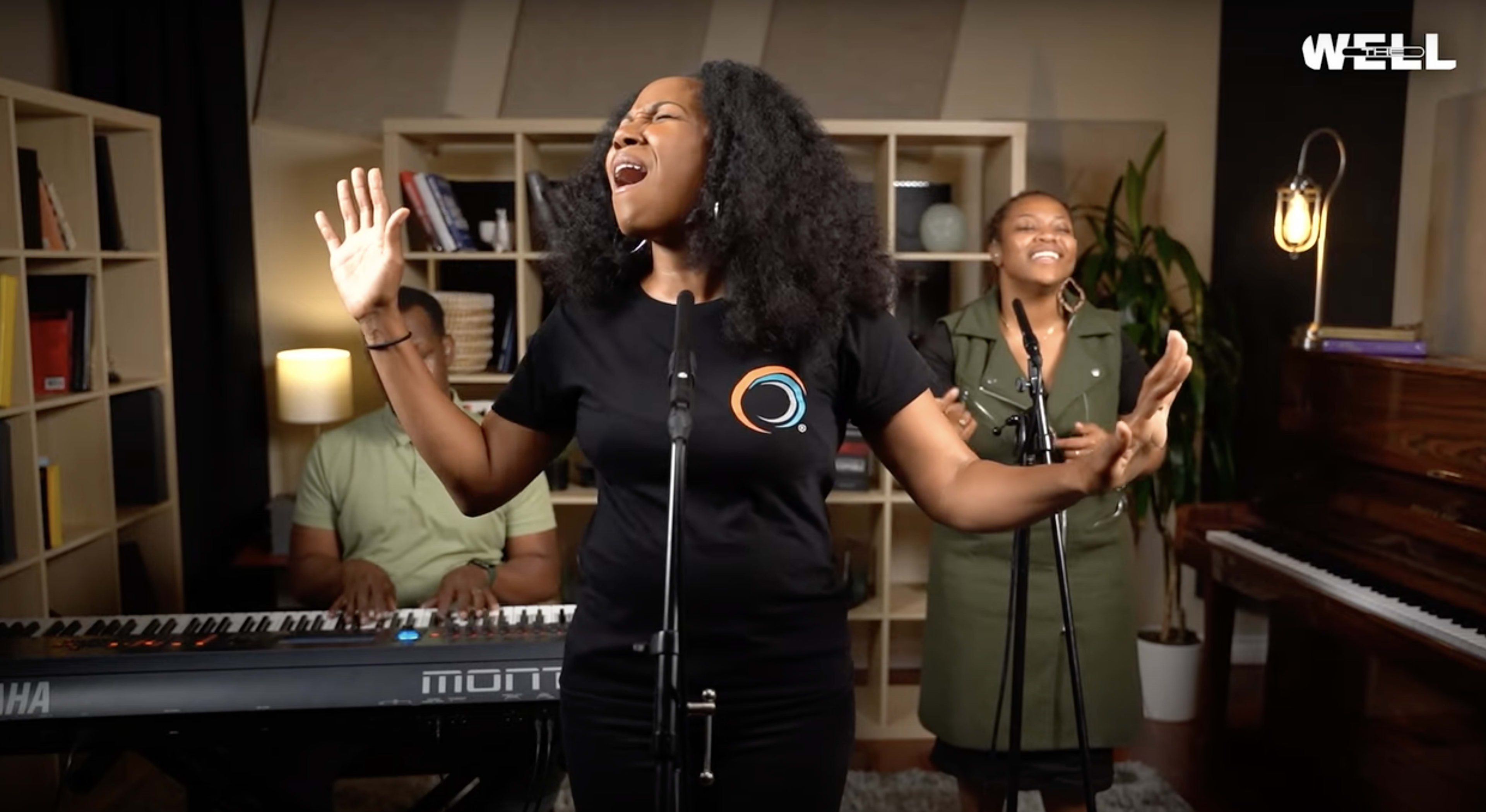 A woman sings passionately on stage while a man plays piano and another woman smiles in the background.