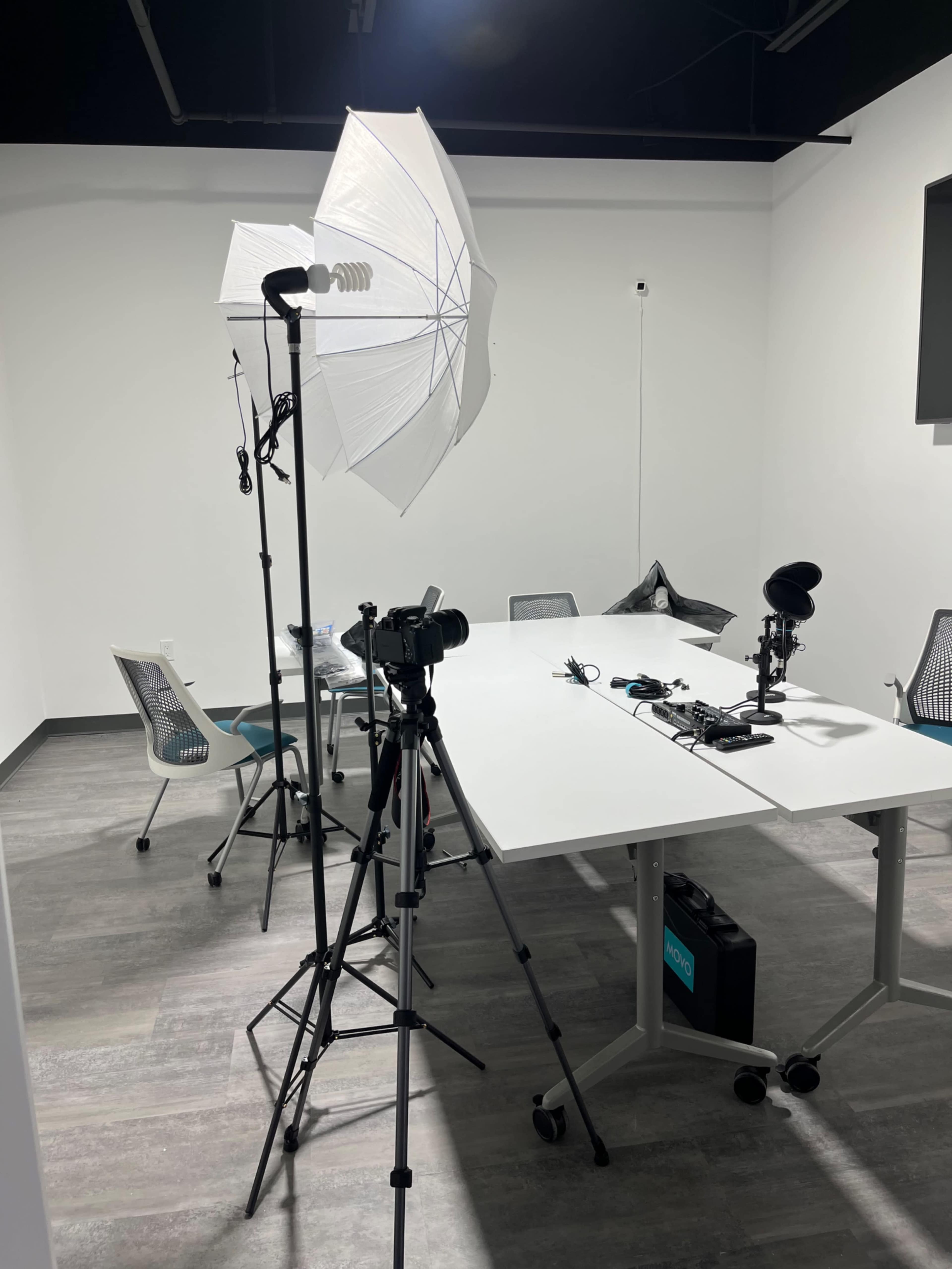 The image shows a well-lit studio setup with a camera on a tripod, lighting equipment, and a table in a modern meeting room.