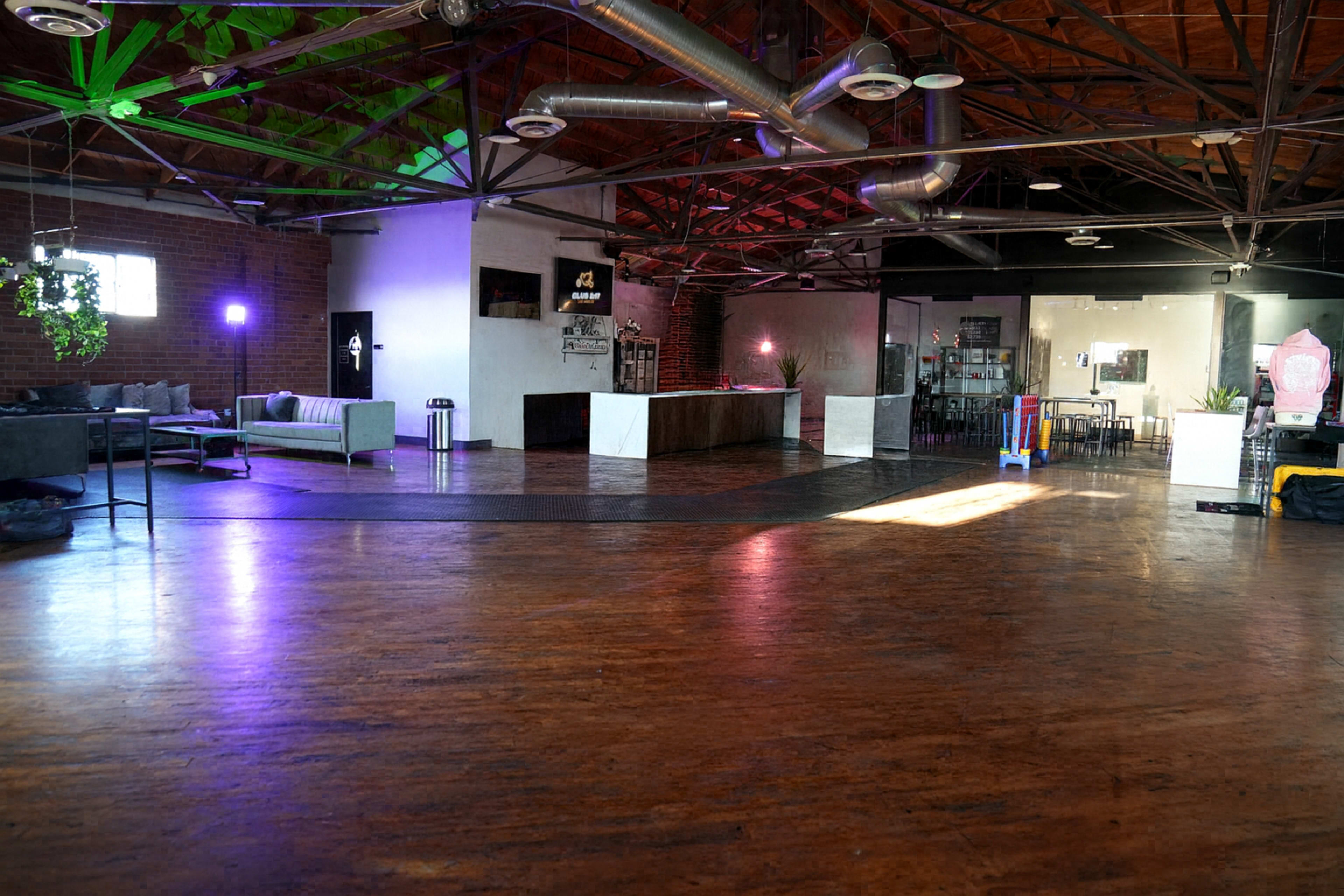 The image shows an open, industrial-style event space with wooden flooring, exposed beams, and modern furnishings, including a seating area and a bar.