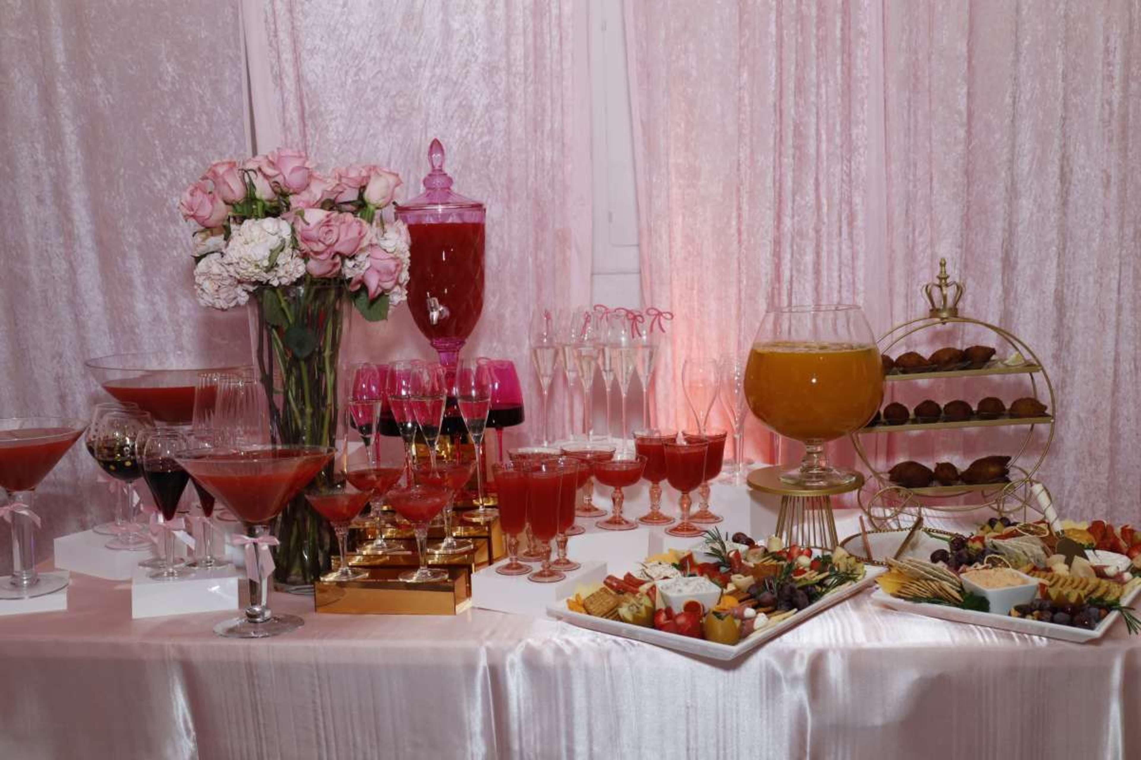 The image shows a festive table set with various colorful cocktails, a floral arrangement, and a selection of appetizers arranged on platters.