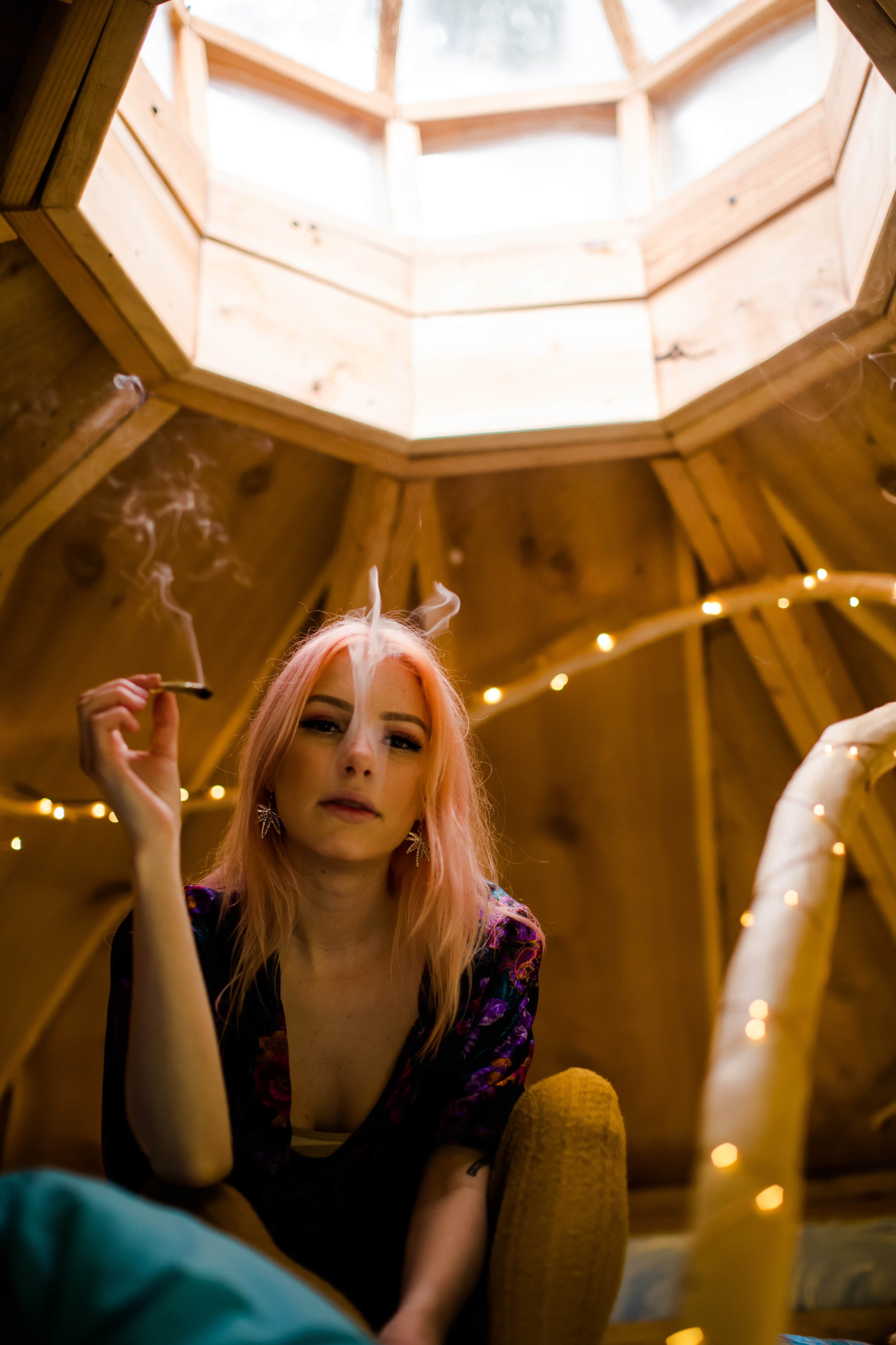 A woman with pink hair sits inside a wooden structure, smoking a cigarette, while warm string lights illuminate the space around her.