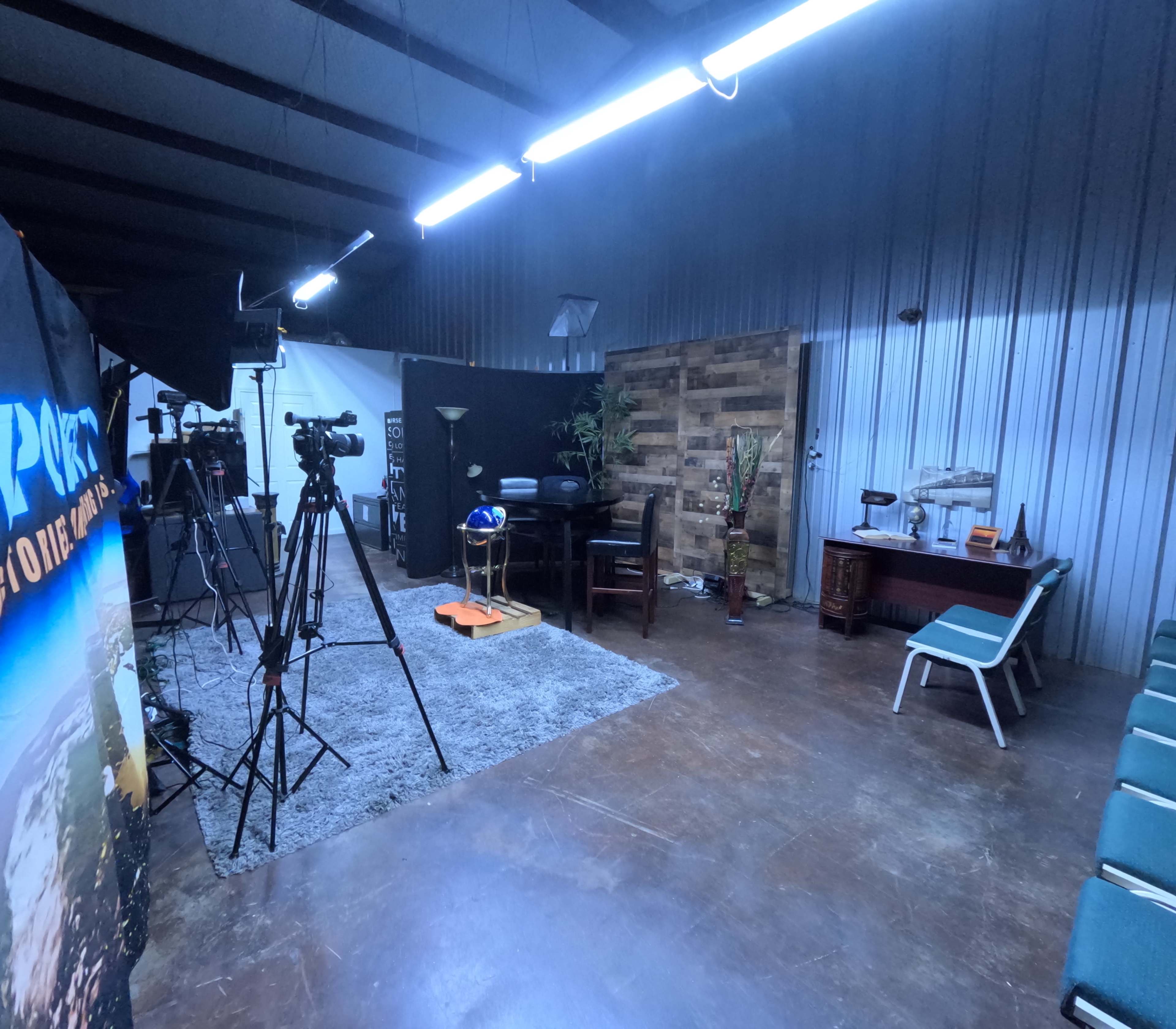 The image shows a studio setup with cameras, lights, and a small seating area, featuring a wooden wall panel and various furnishings.