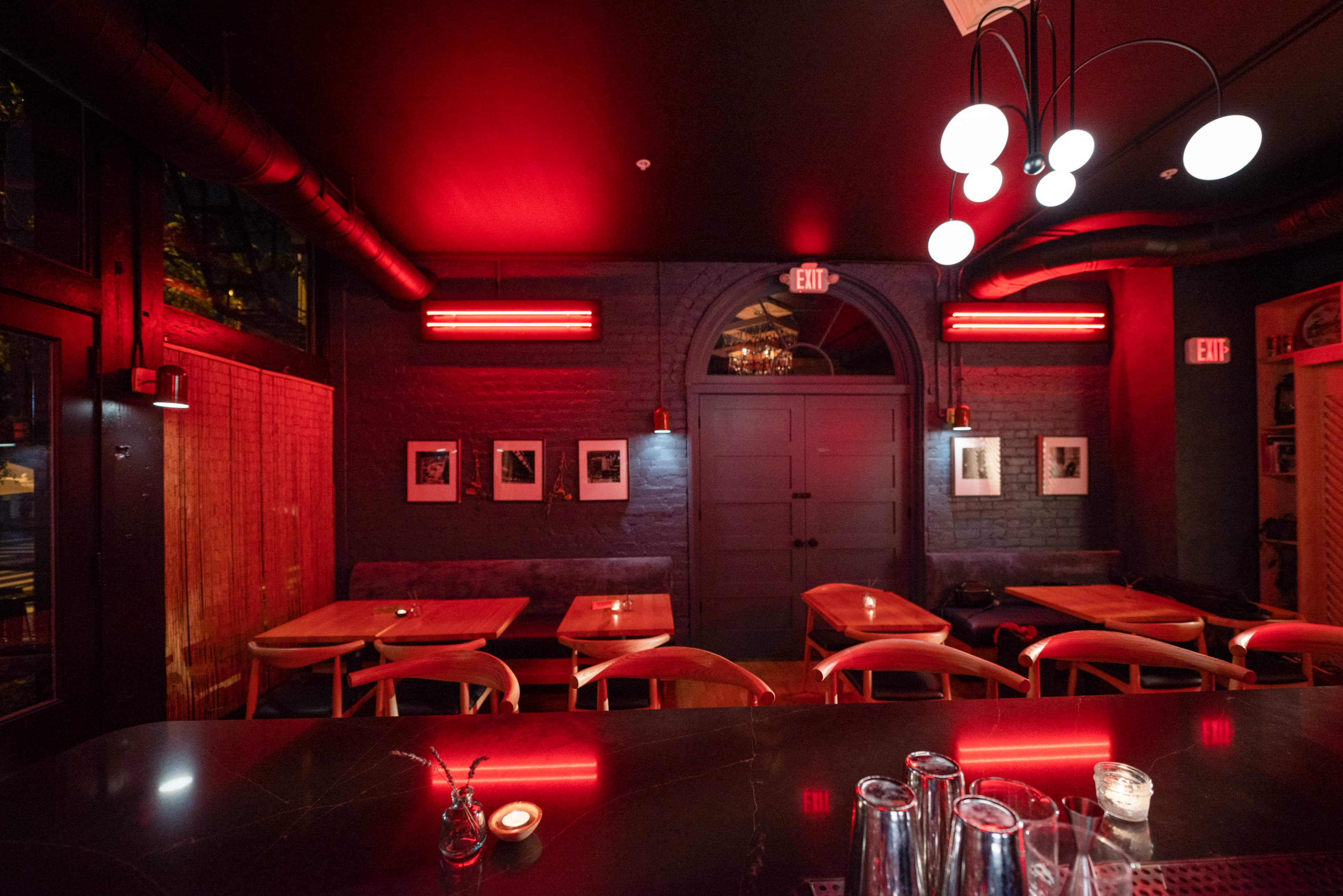 A dimly lit restaurant interior with red lighting, featuring wooden tables, framed photographs on the walls, and a bar area with glasses.