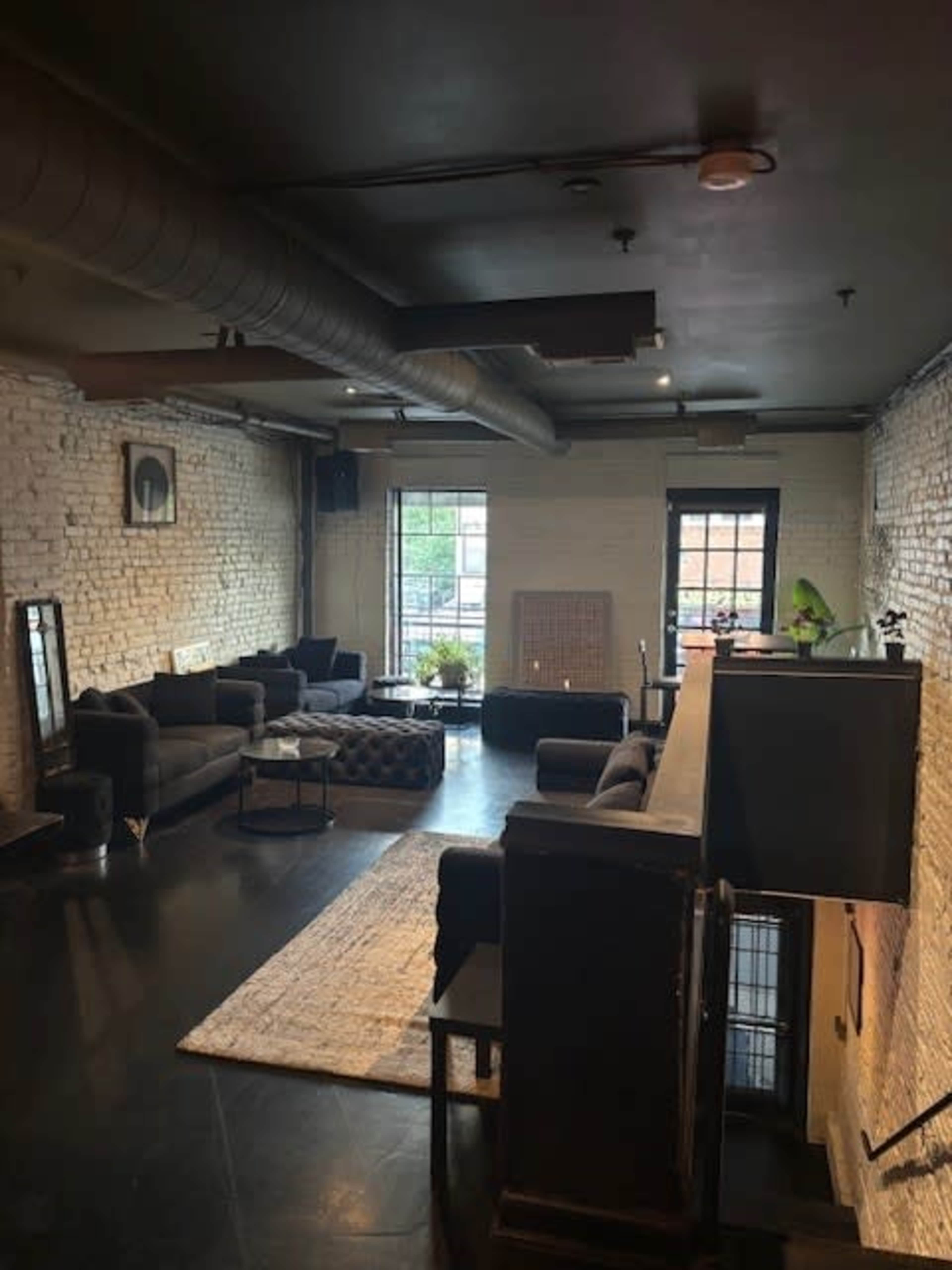 The image shows a spacious, modern lounge area with gray sofas, a patterned ottoman, and large windows letting in natural light.