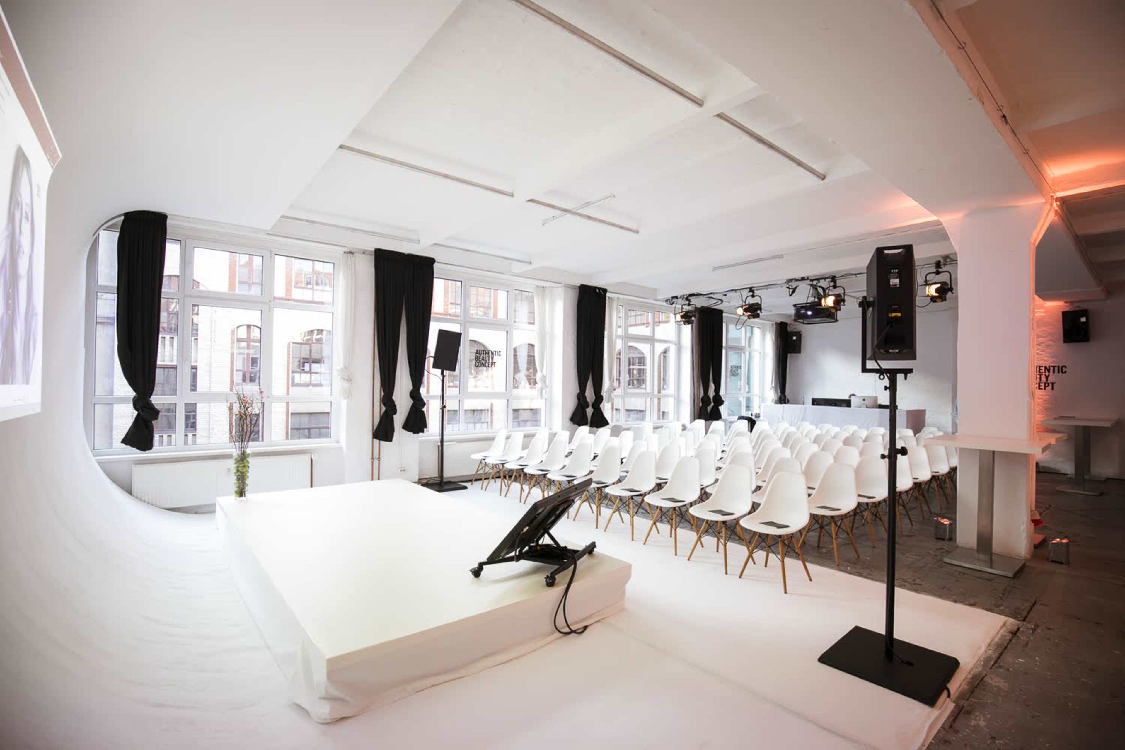 A minimalist event space set up with a stage, a row of white chairs, and large windows letting in natural light.