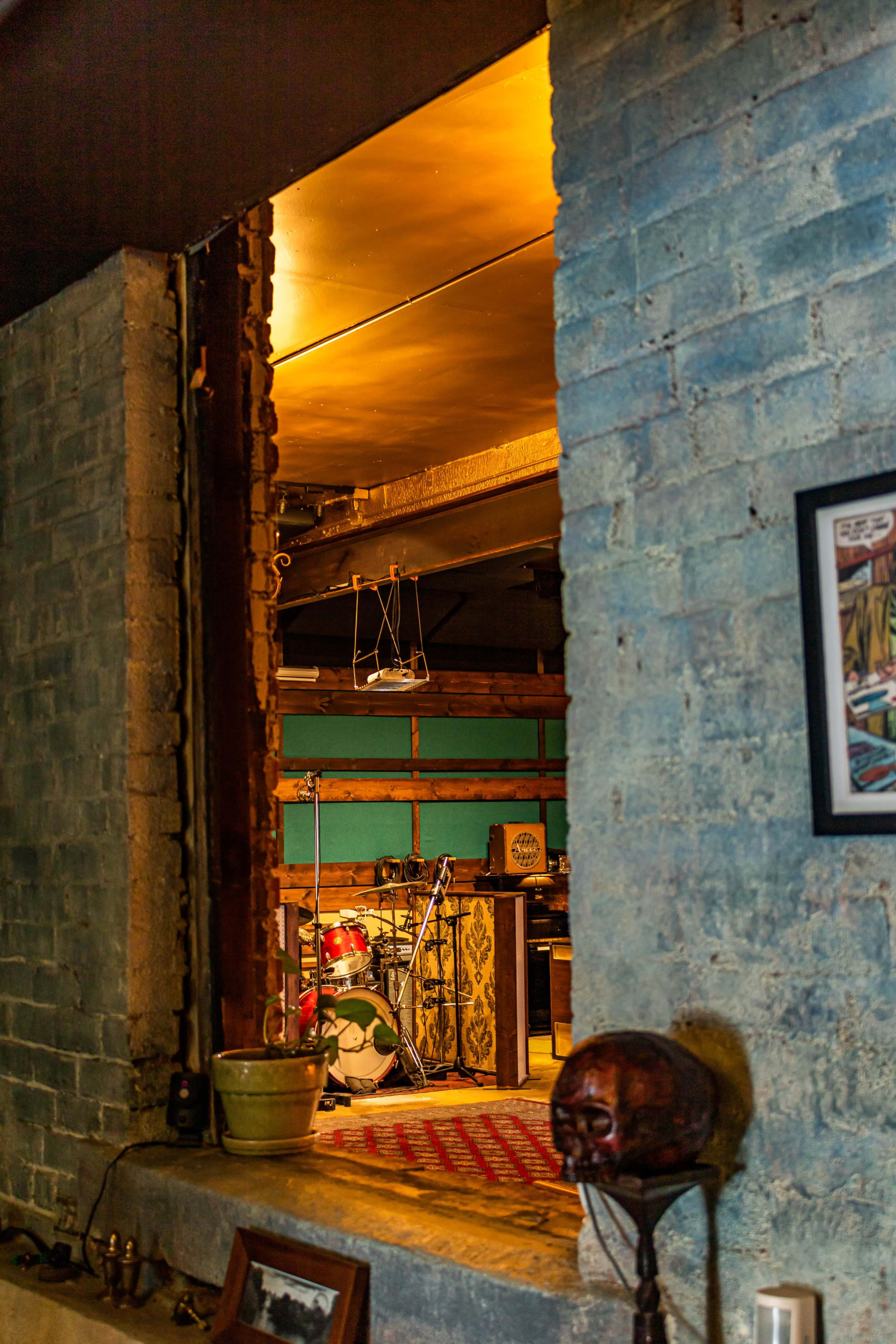 A view through a partially completed brick wall reveals a room with a drum set and various furnishings.