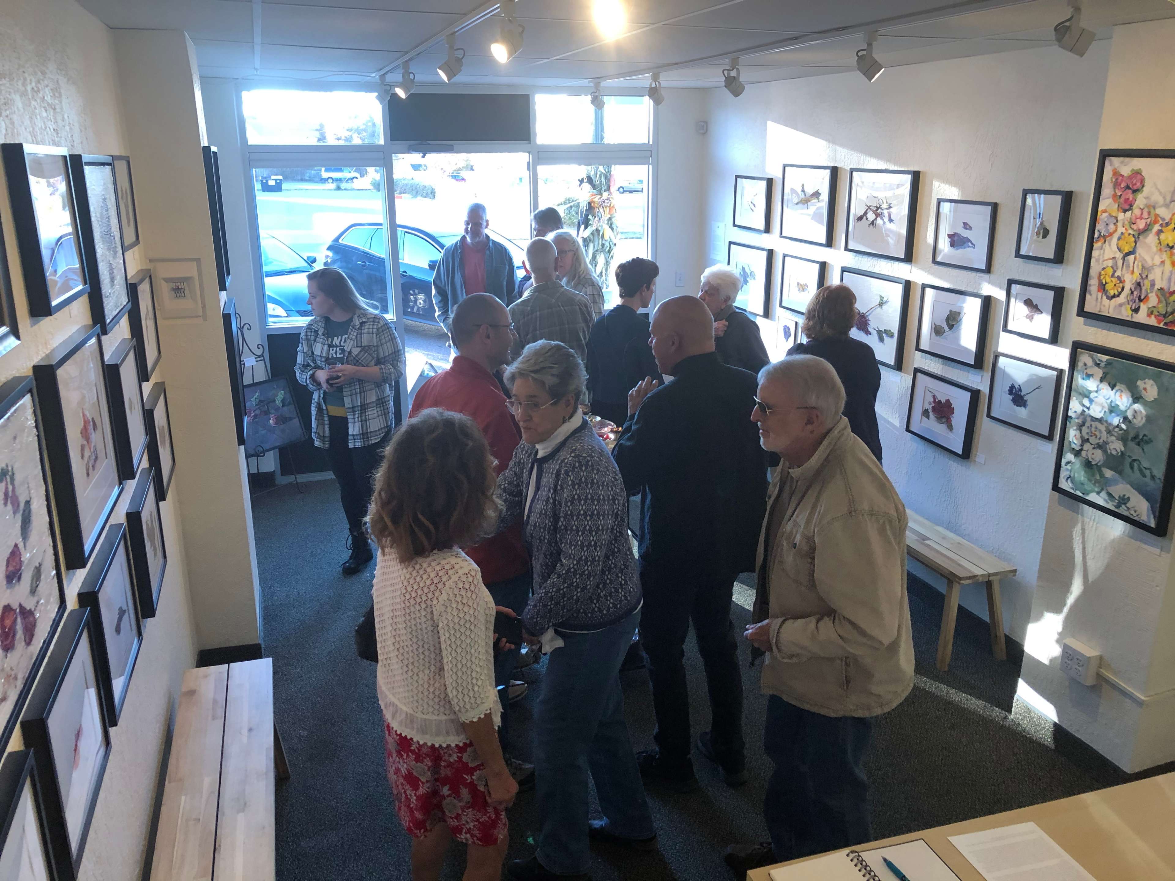 A group of people stands inside an art gallery, surrounded by framed artworks on the walls, with some conversing and others observing the pieces.