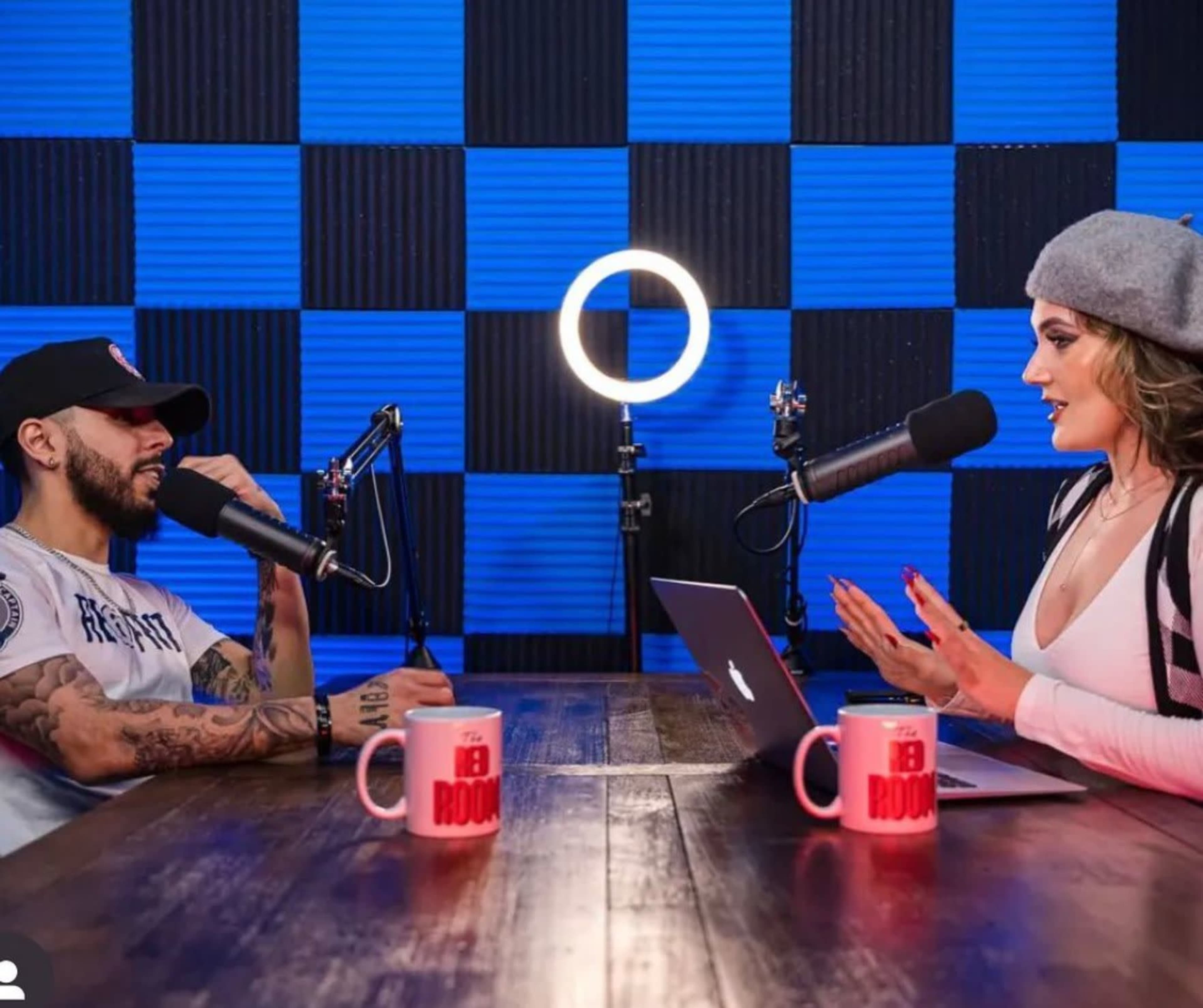 A man and a woman are seated at a table with microphones, engaged in a discussion, in front of a backdrop of blue and black checkered panels.