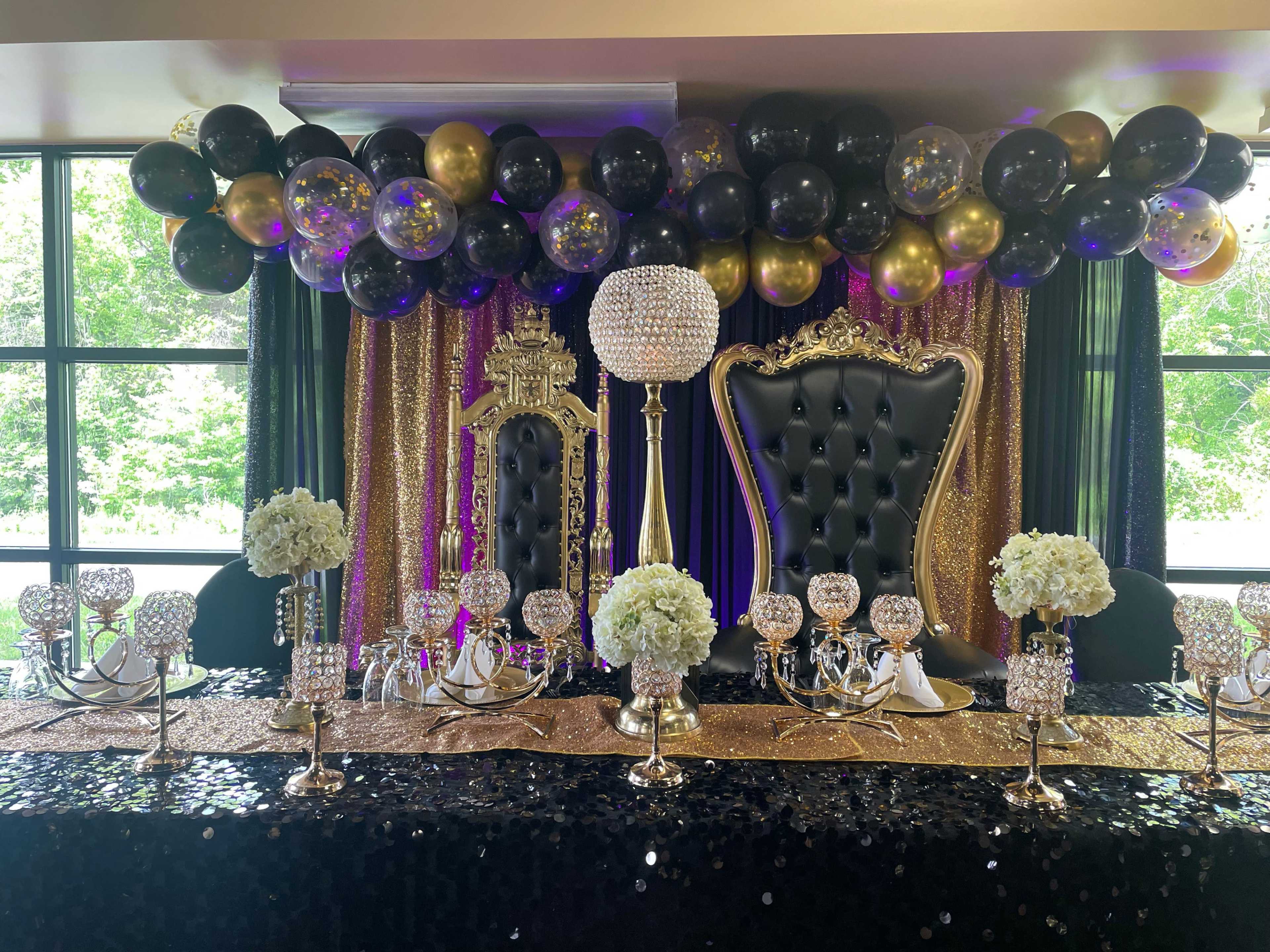 An elegantly decorated event table featuring two large thrones, a display of flowers, and a backdrop of black, gold, and purple balloons.