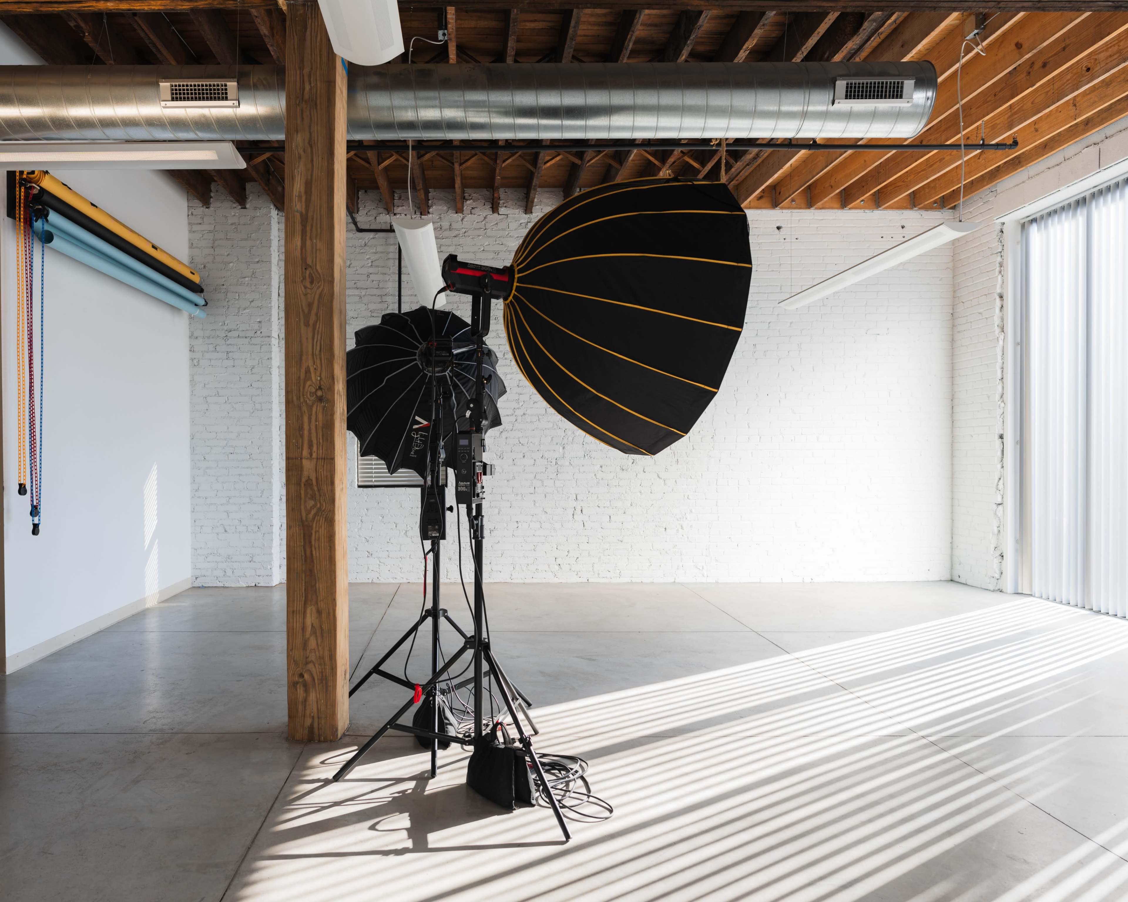 The image shows a photography studio featuring a backdrop of white brick walls and concrete flooring, with lighting equipment set up on stands.