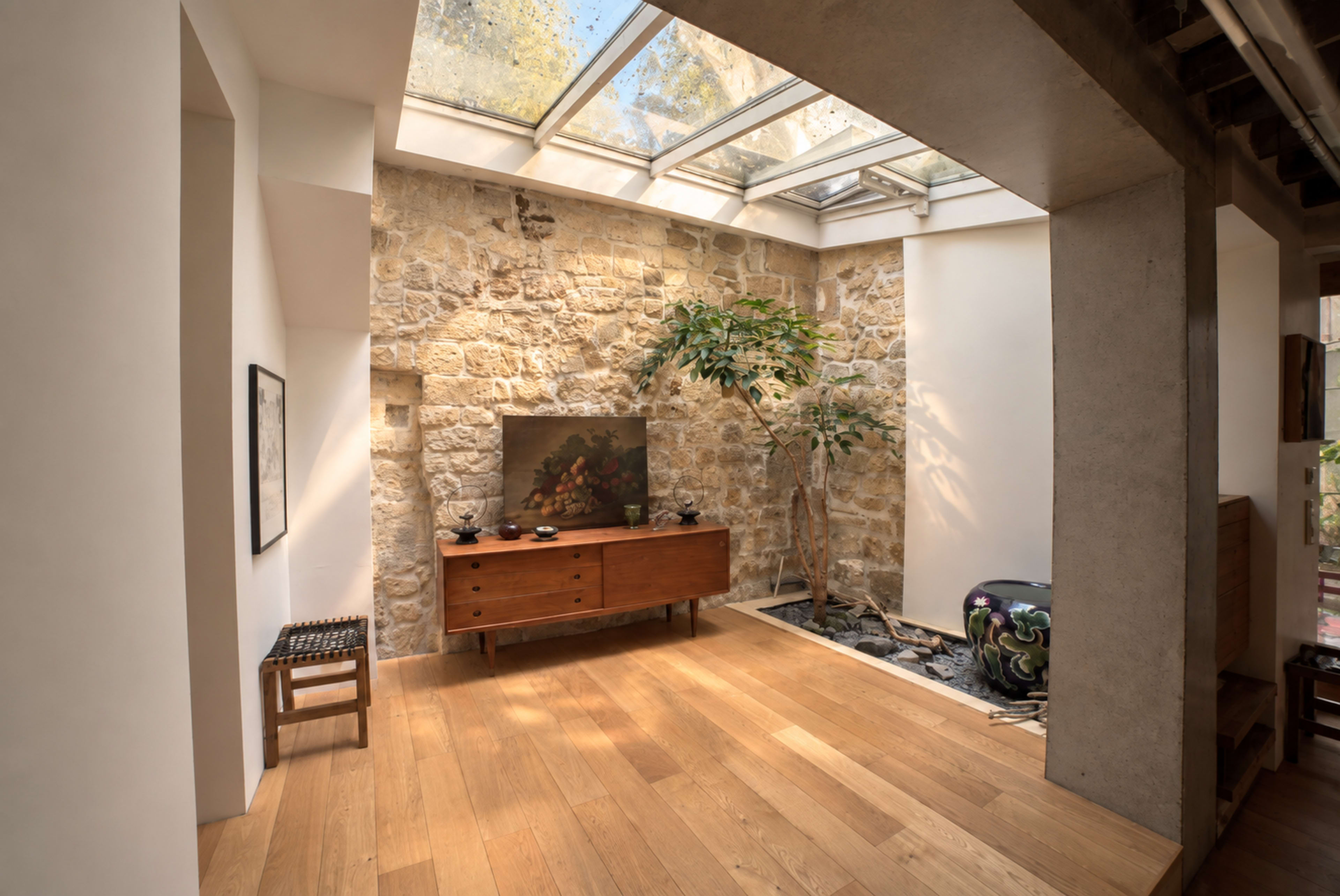 The image shows a well-lit interior space with a stone wall, wooden flooring, and a large window above, featuring a plant and a decorative painting on a wooden sideboard.