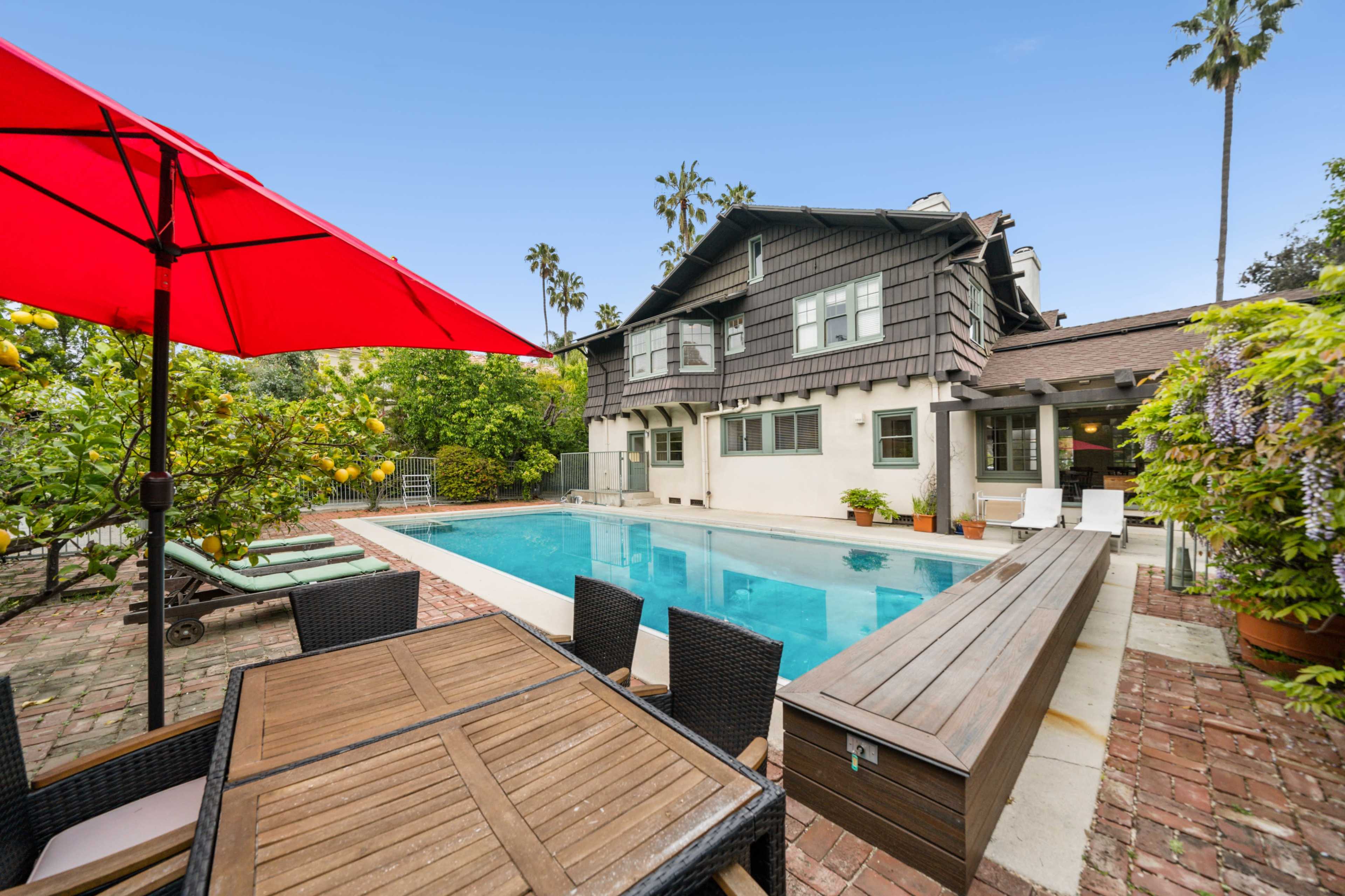 A backyard with a swimming pool, a dining table, and a house surrounded by greenery and palm trees.