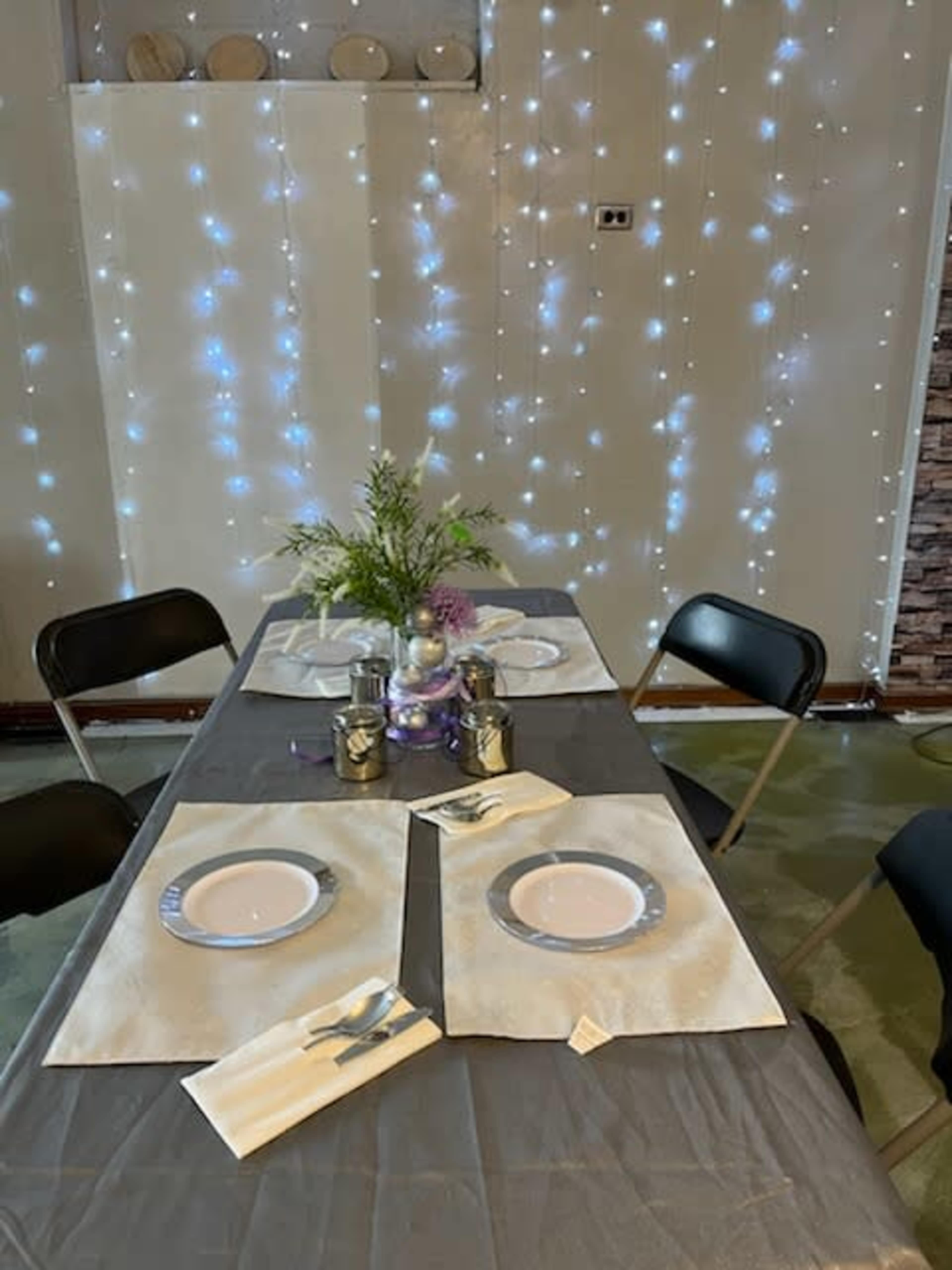 A dining table is set with four plates and decorative elements, surrounded by chairs and illuminated by strands of twinkling lights on the wall.