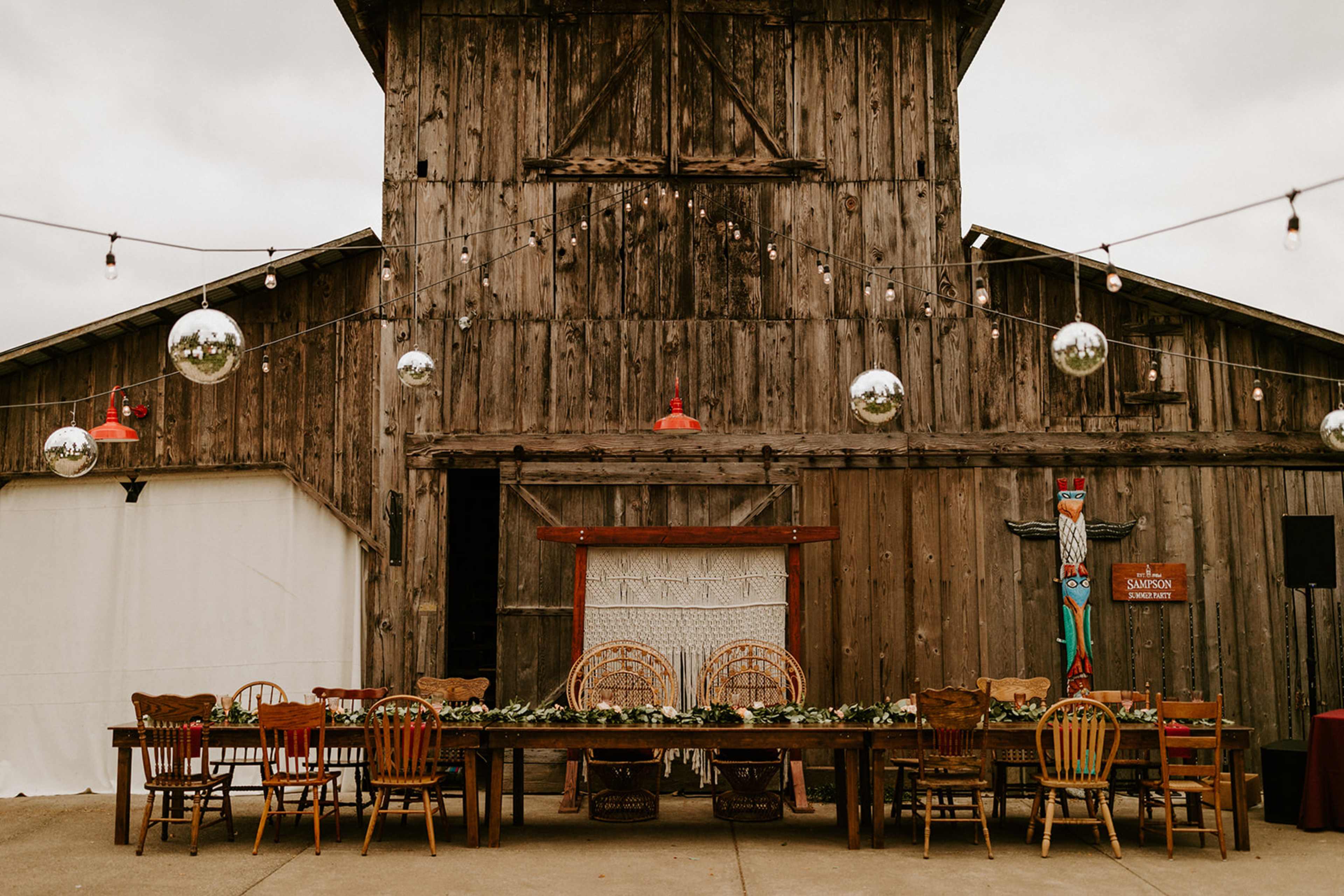 A long dining table is set in front of a rustic wooden barn, adorned with hanging disco balls and string lights.