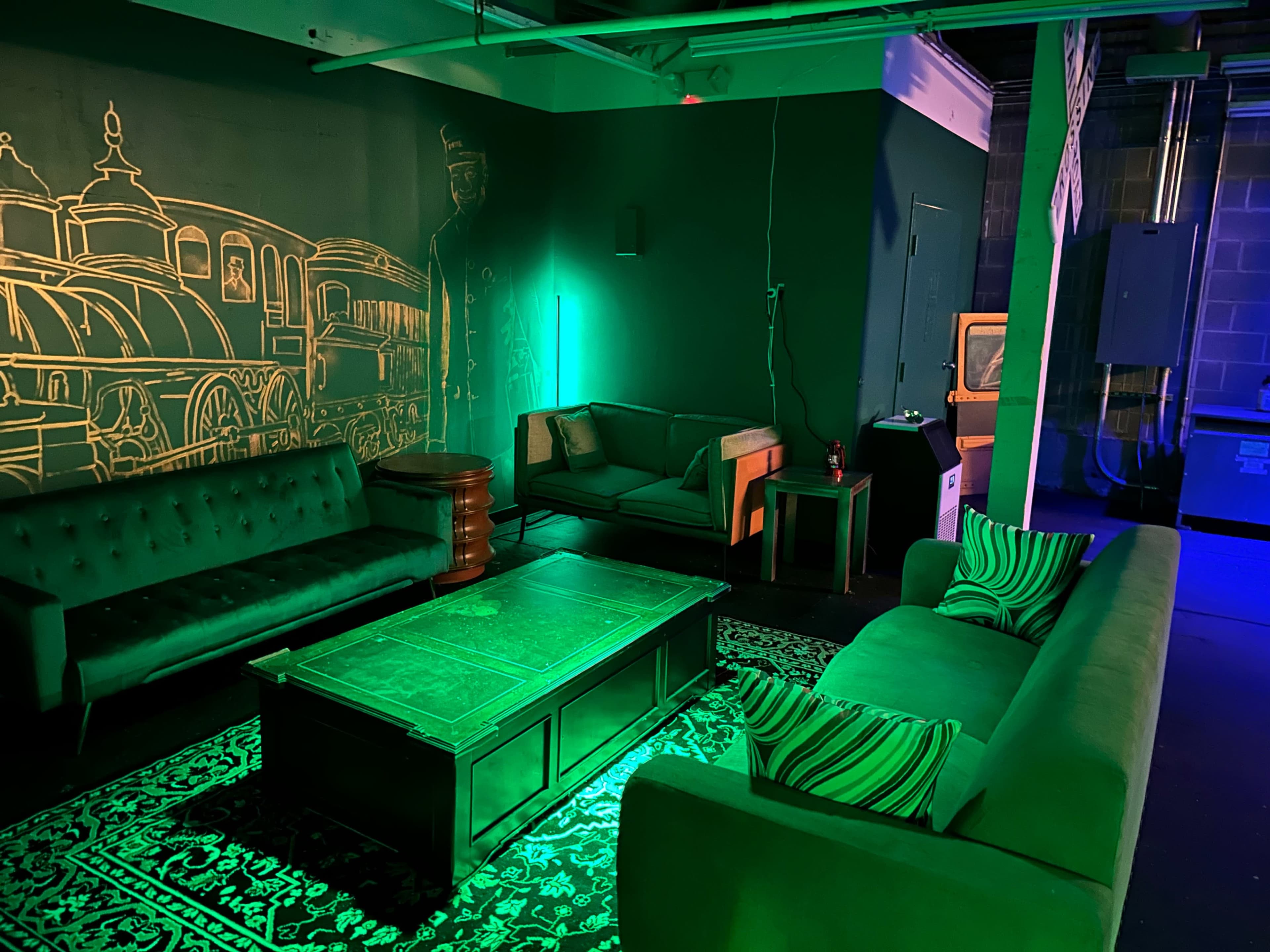 A lounge area with green lighting, a dark wall mural of a train, two sofas, a coffee table, and a patterned rug.