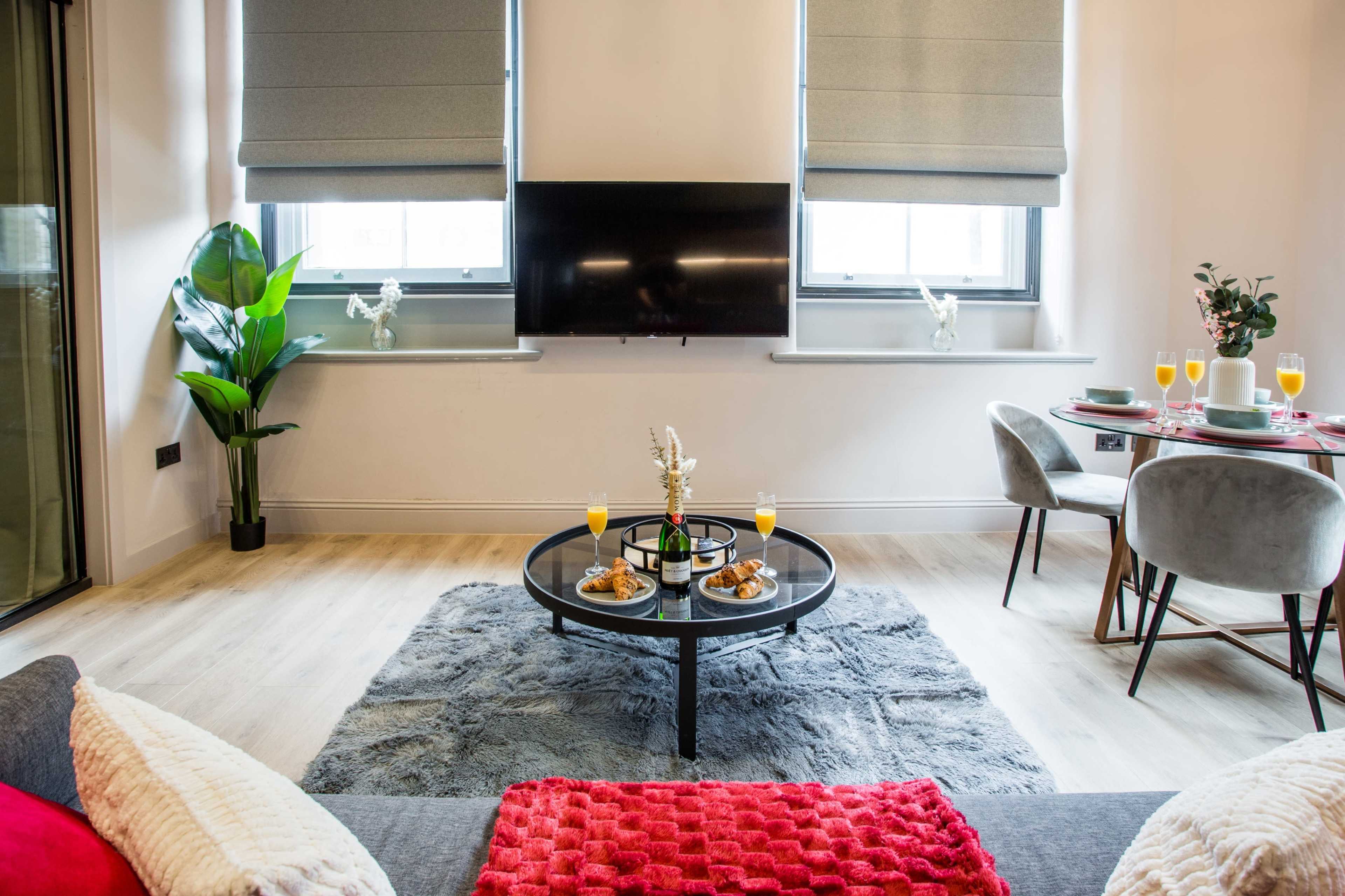The image shows a modern living room with a coffee table set for breakfast, featuring orange juice and pastries, and a dining area with two chairs.