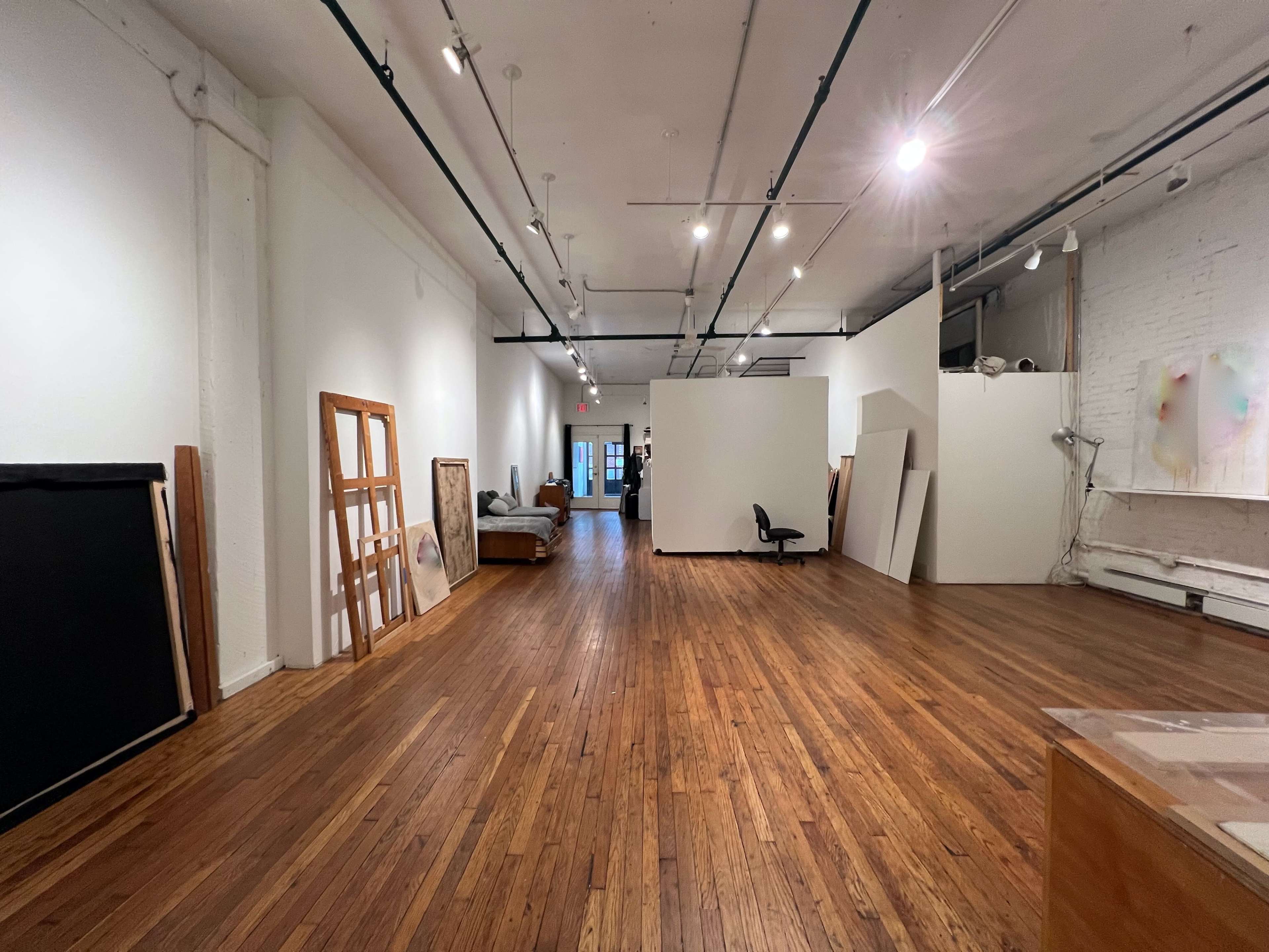 A spacious gallery with wooden flooring, white walls, and several pieces of art and furniture leaning against the walls.