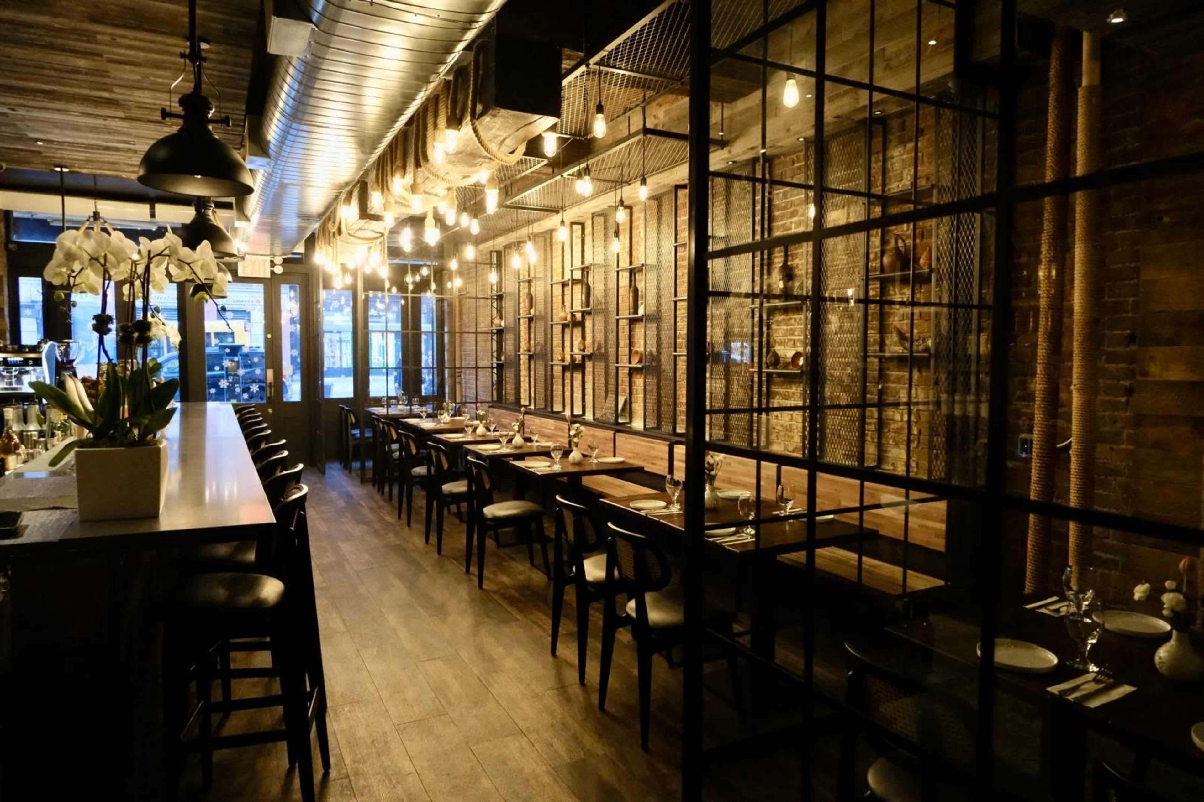 The image shows a dimly lit restaurant interior featuring wooden walls, a long dining table with chairs, and decorative lighting.