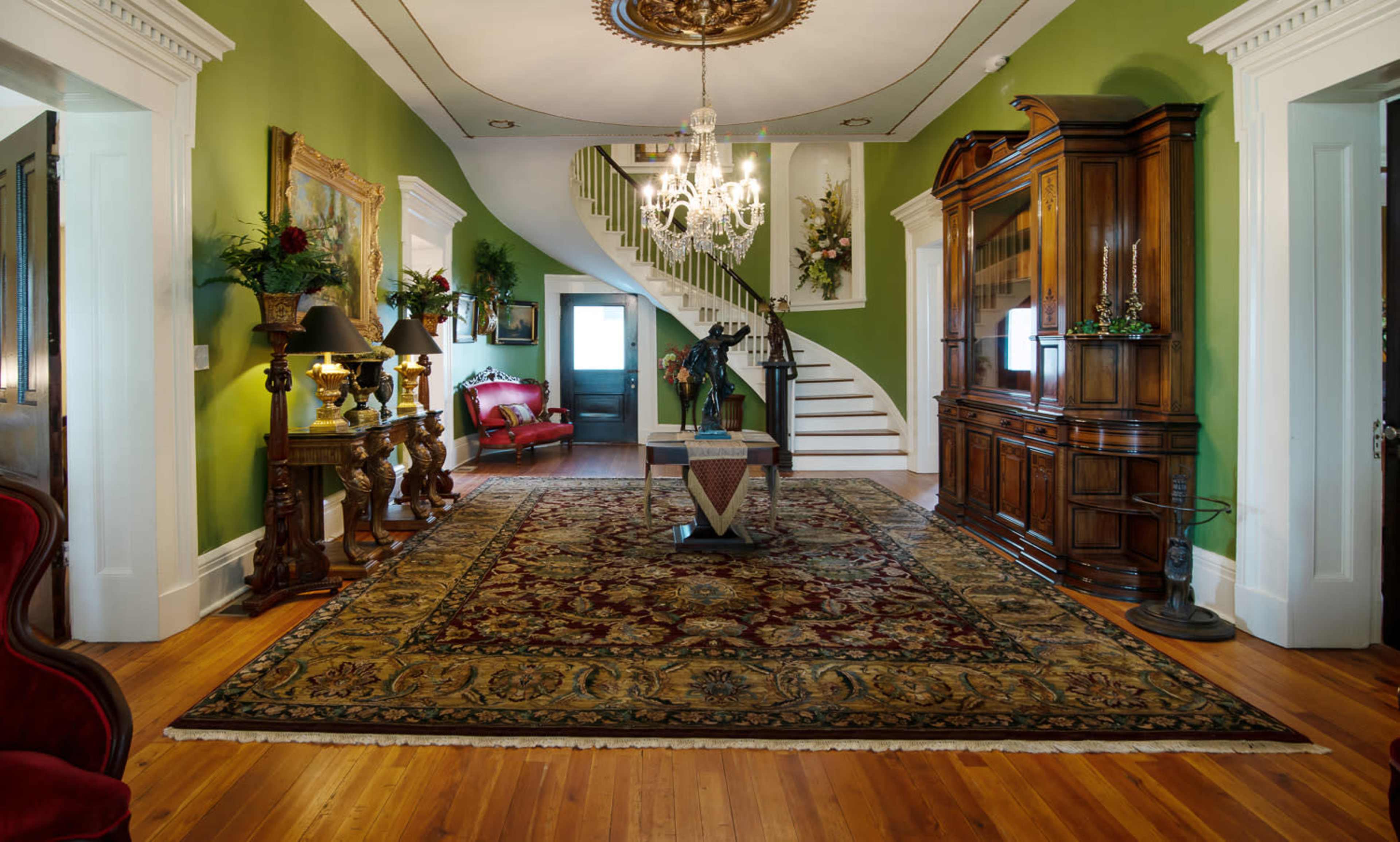 A foyer with green walls, a chandelier, a staircase, and wooden furniture, including a decorative cabinet and a large area rug.