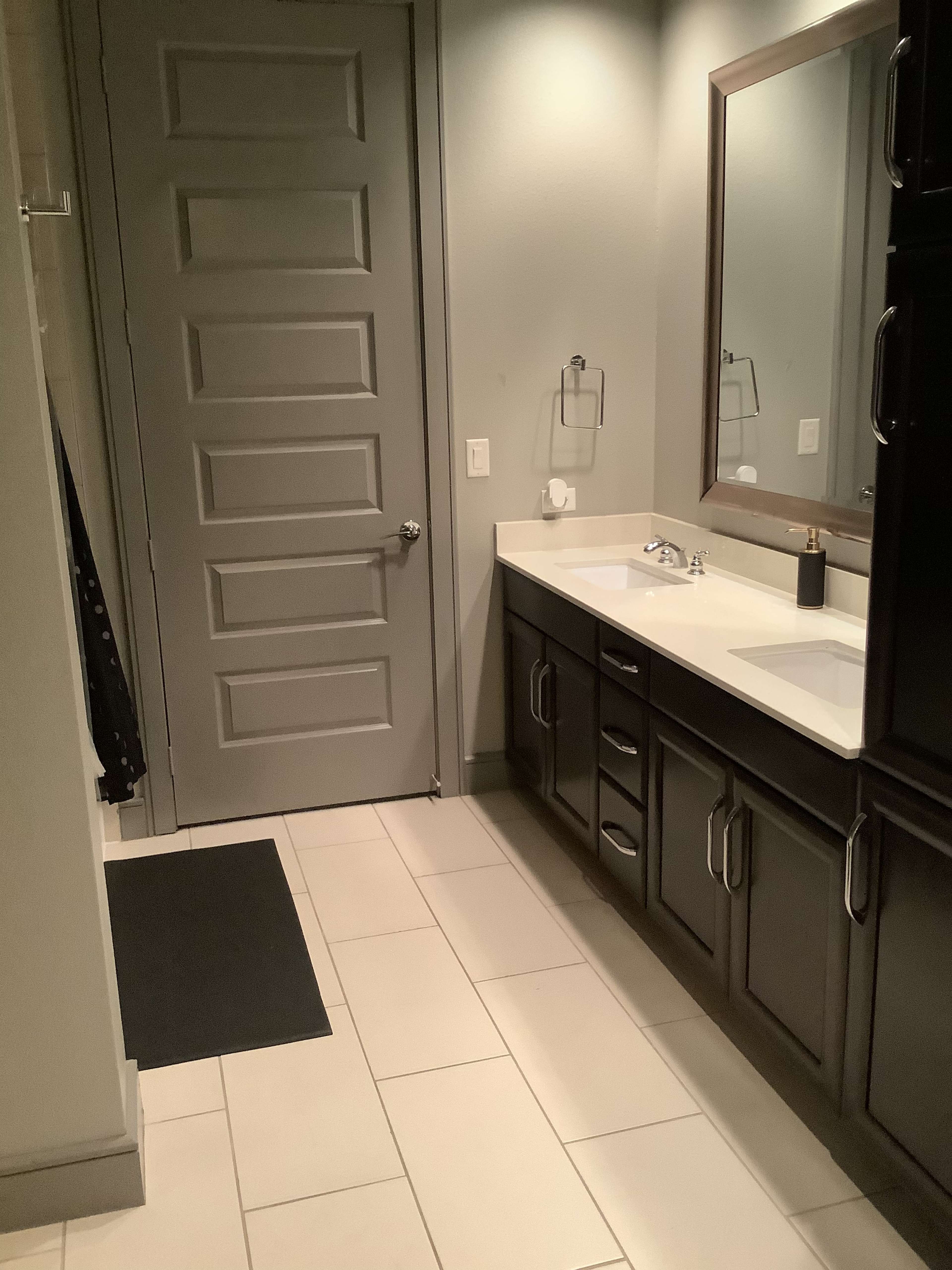 A modern bathroom features a double-sink vanity, a large mirror, and a door leading to another area.