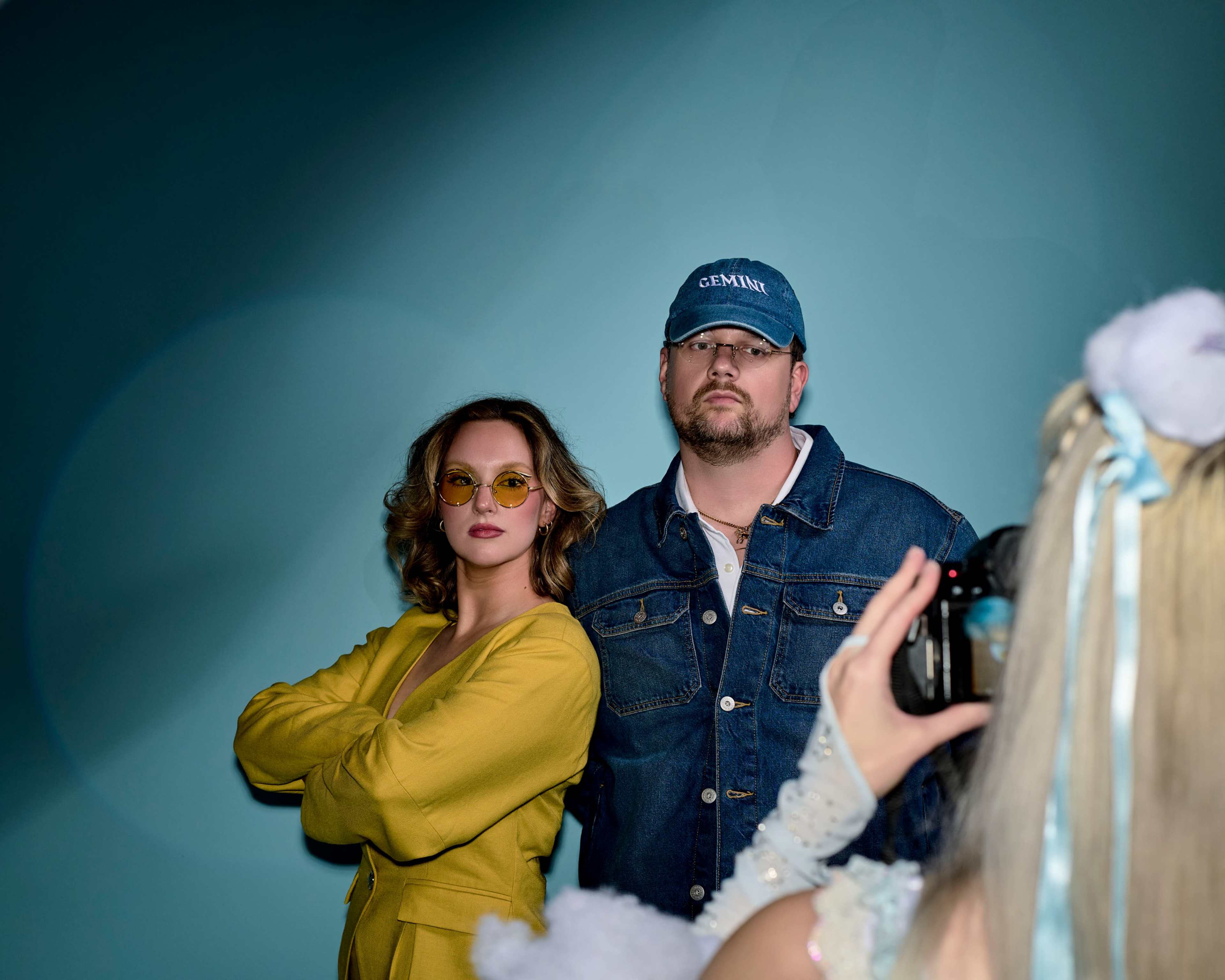 A woman in yellow attire and round sunglasses stands confidently next to a man in a blue cap and denim jacket, while another person is taking their picture.
