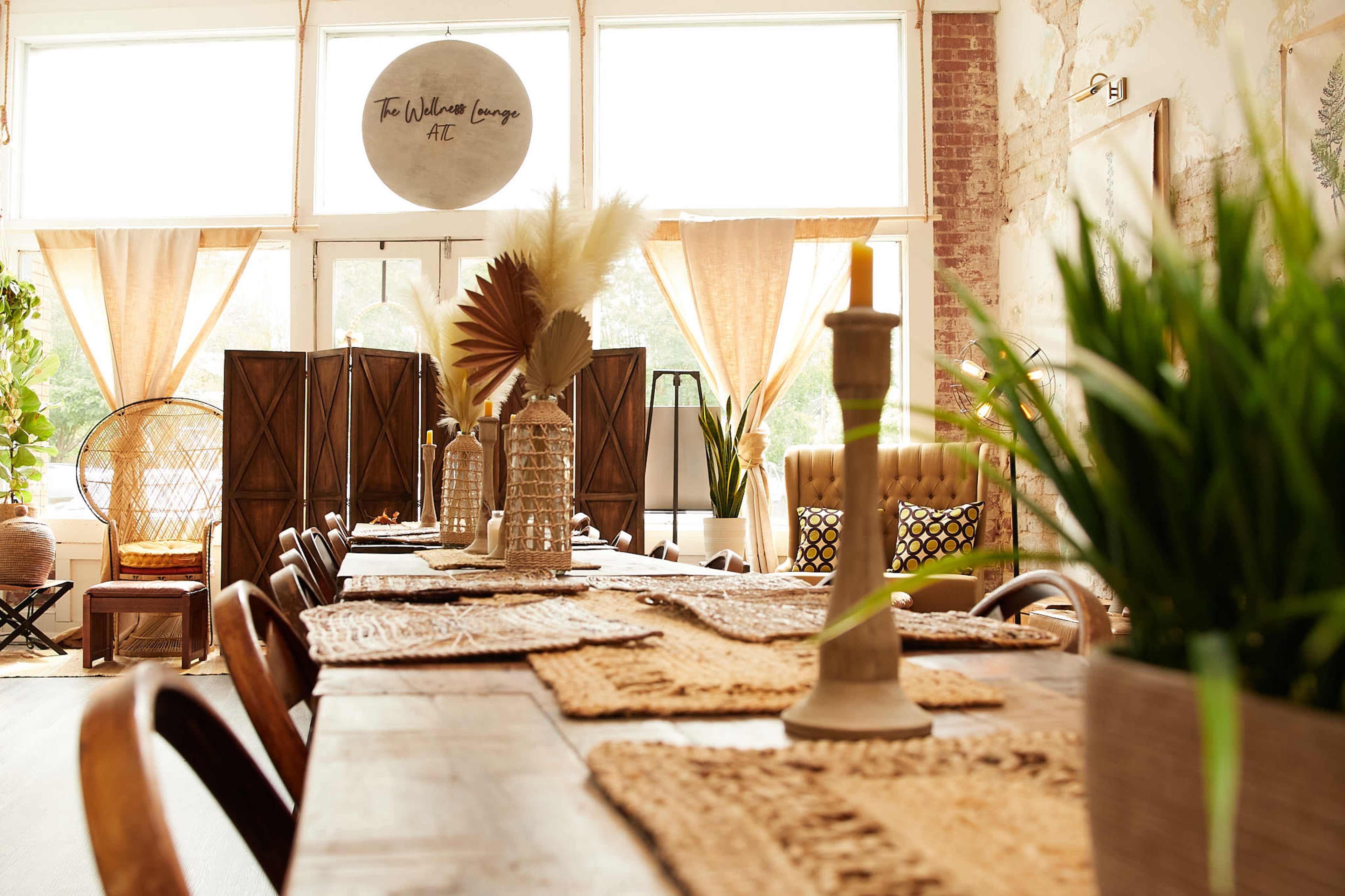 A long wooden dining table is set in a brightly lit room decorated with plants, woven decor, and cozy seating arrangements.