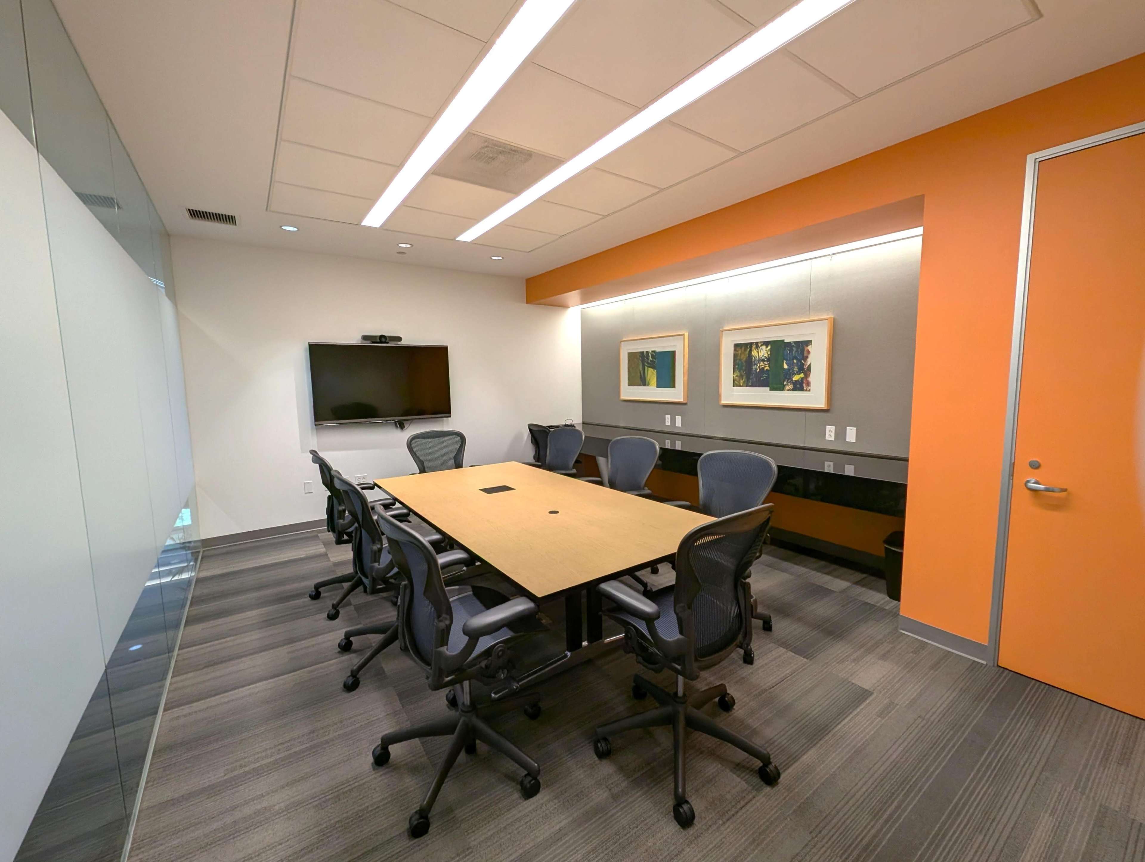 The image shows a modern conference room with a large rectangular table surrounded by ergonomic chairs, featuring an orange accent wall and a mounted television.