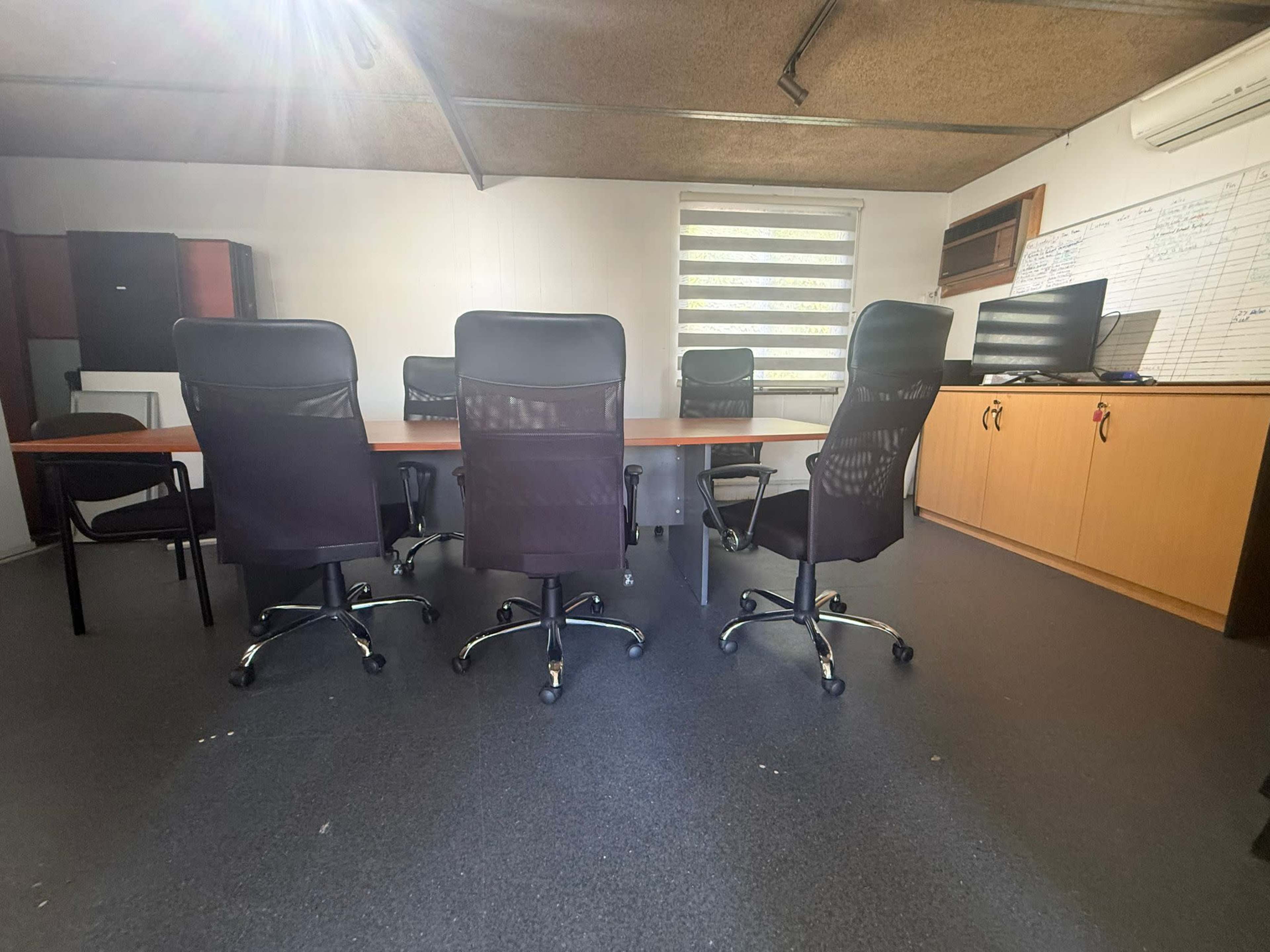 A conference room features a long table with several office chairs arranged around it, along with a monitor and cabinetry in the background.