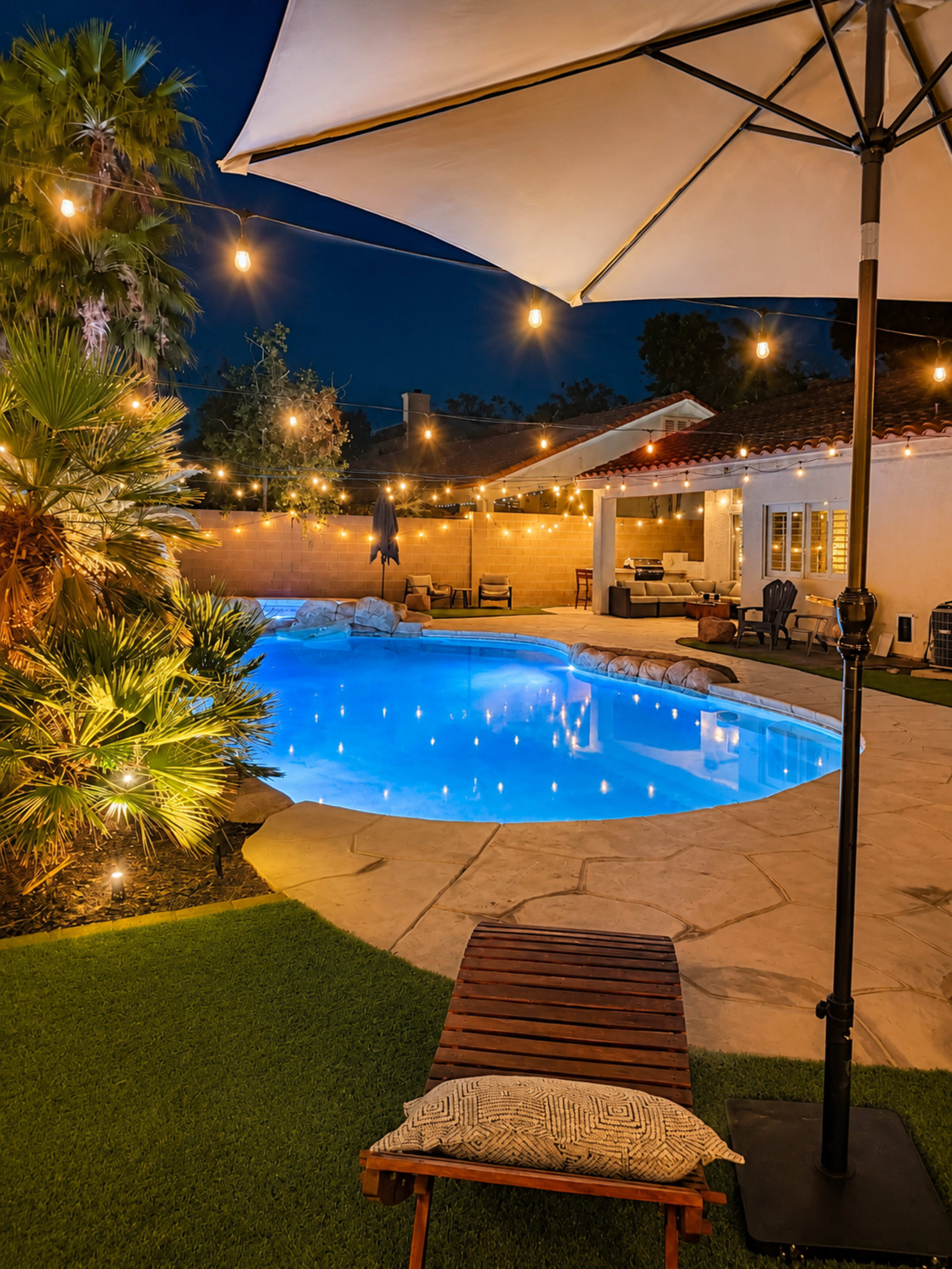 The image shows a illuminated backyard pool area at night, featuring a lounge chair, palm trees, and string lights overhead.