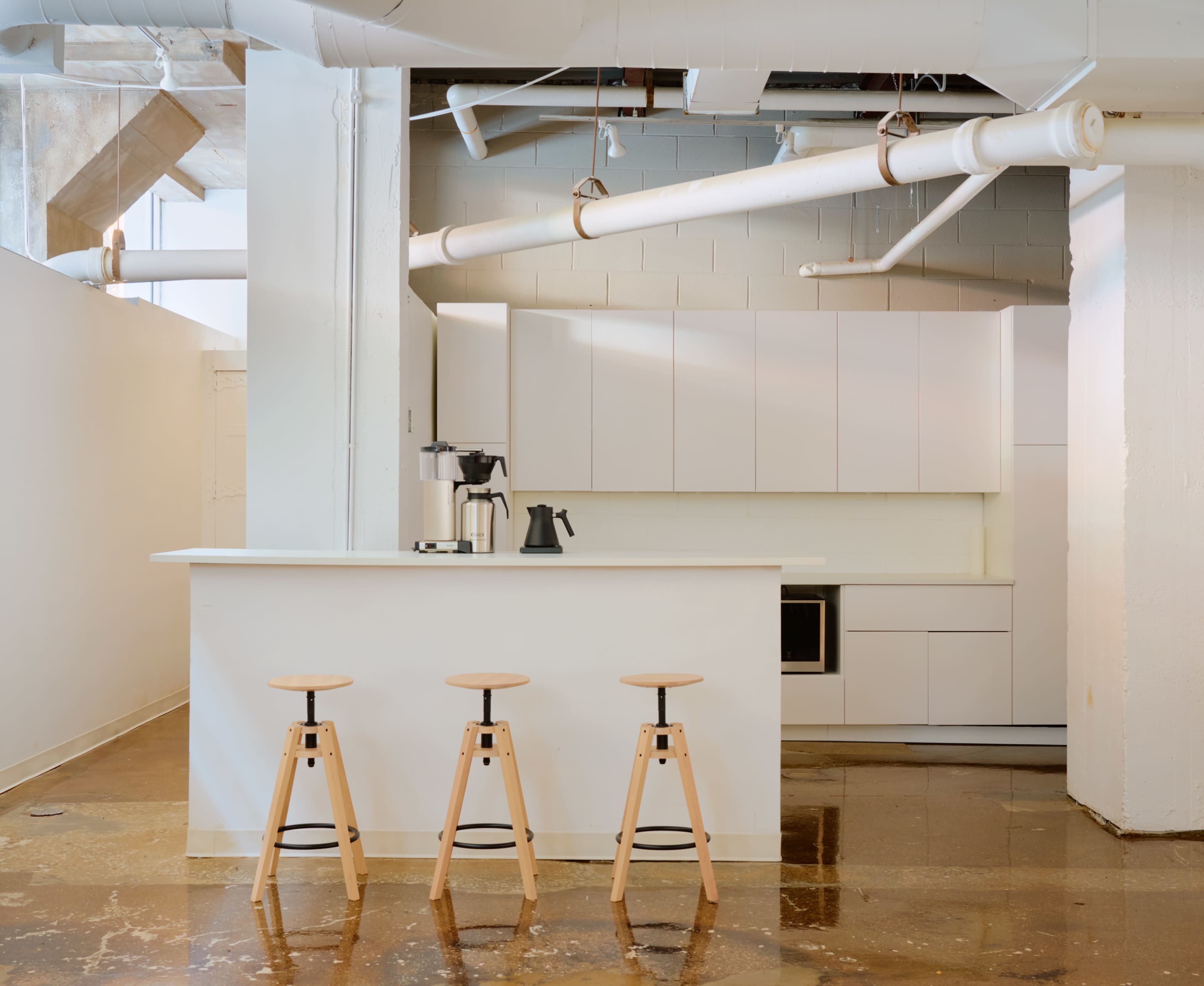 The image features a minimalist kitchen with white cabinetry, a counter with three wooden stools, and exposed plumbing overhead.