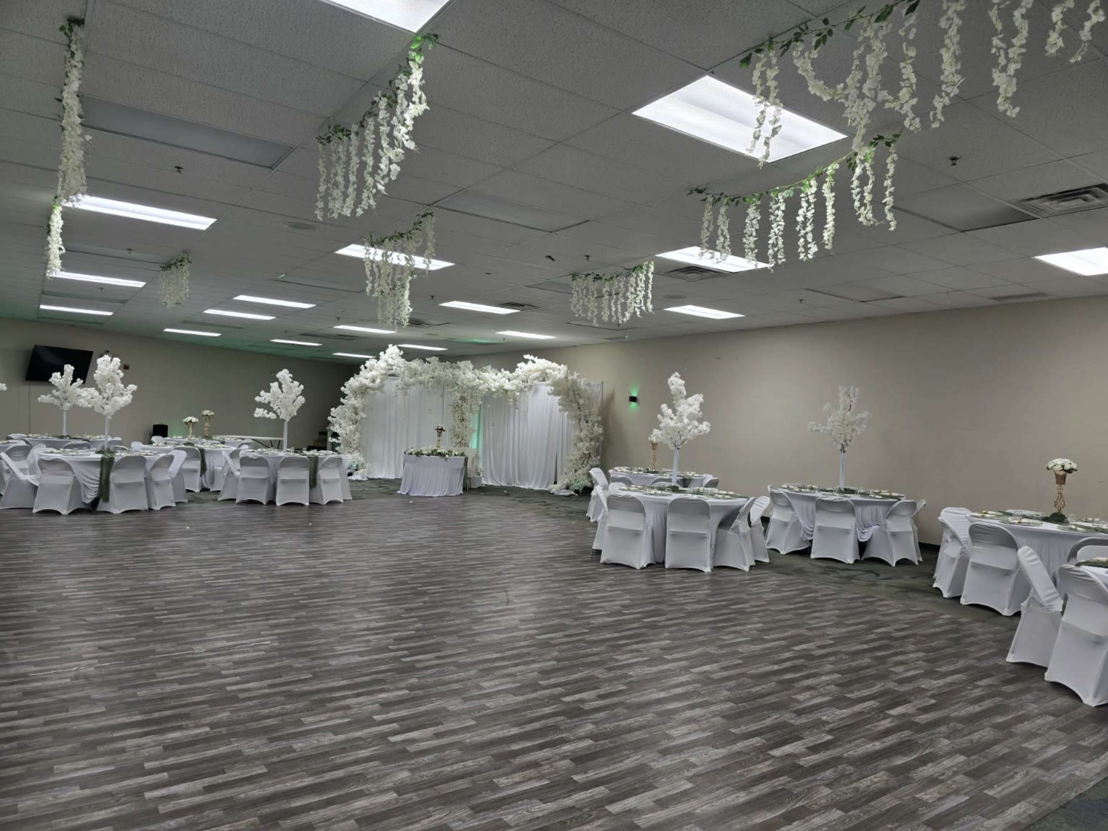 The image shows an indoor event space decorated with white flowers, tables covered with white linens, and a backdrop featuring floral arrangements.