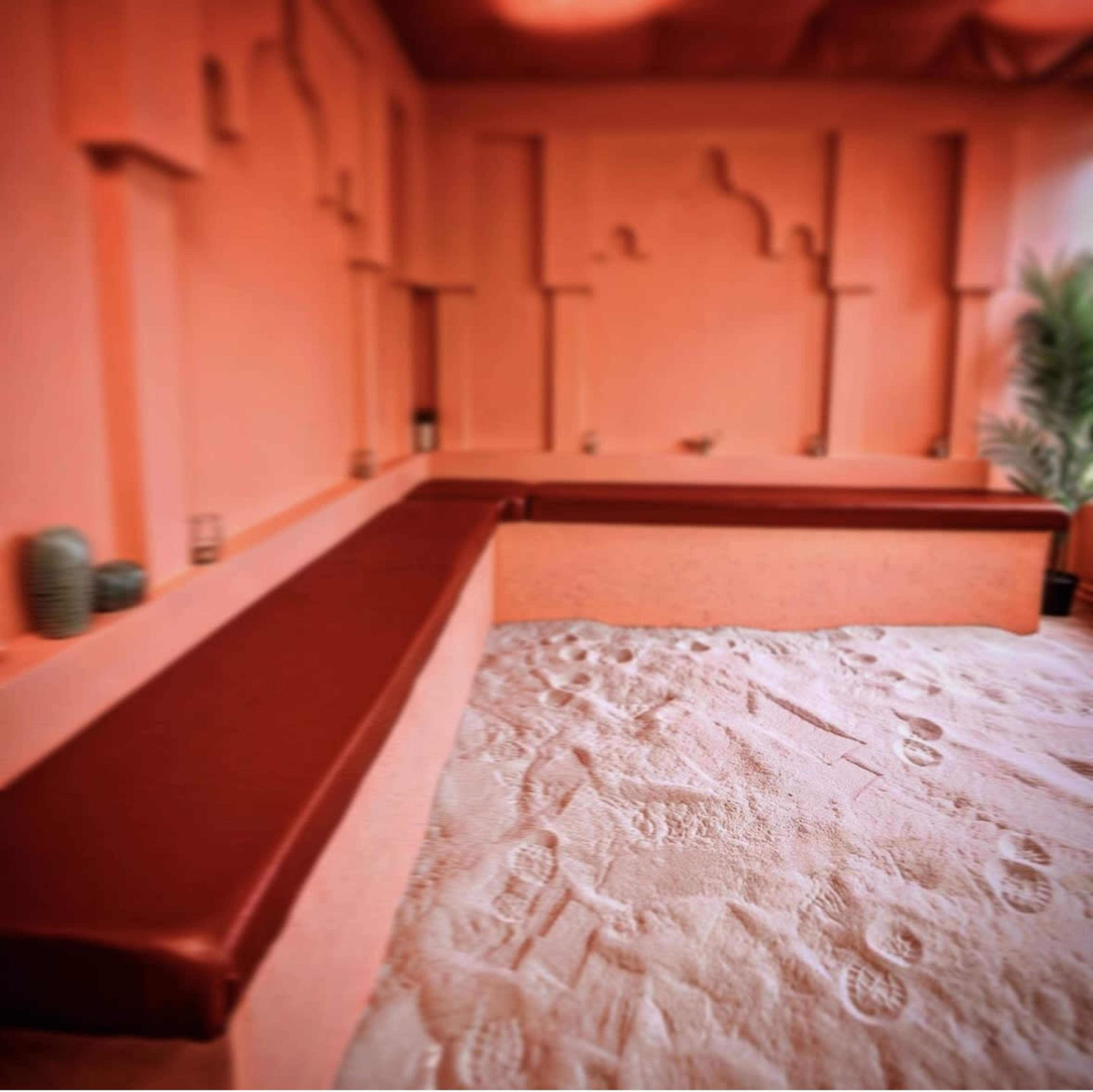 A room with peach-colored walls and a sandy floor, featuring a long brown bench along one side.