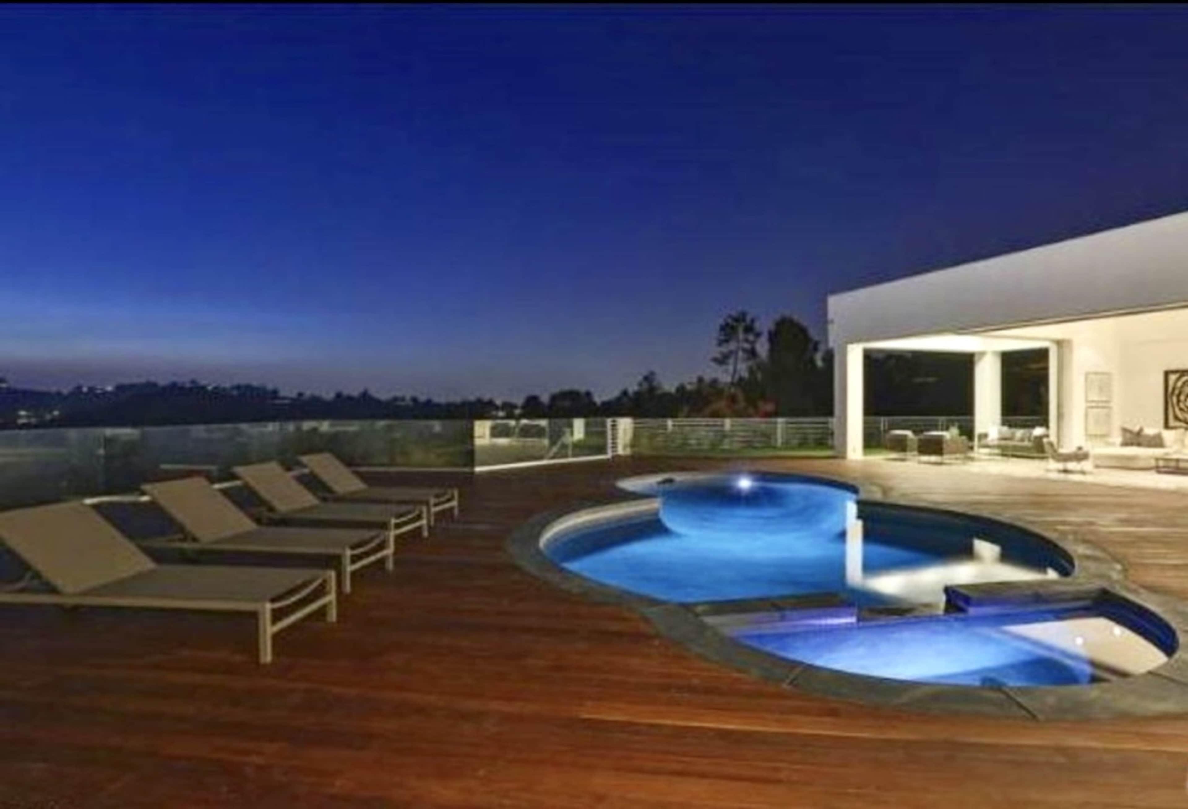 A modern outdoor space featuring a curved swimming pool and lounge chairs on a wooden deck, with a view of the evening sky.