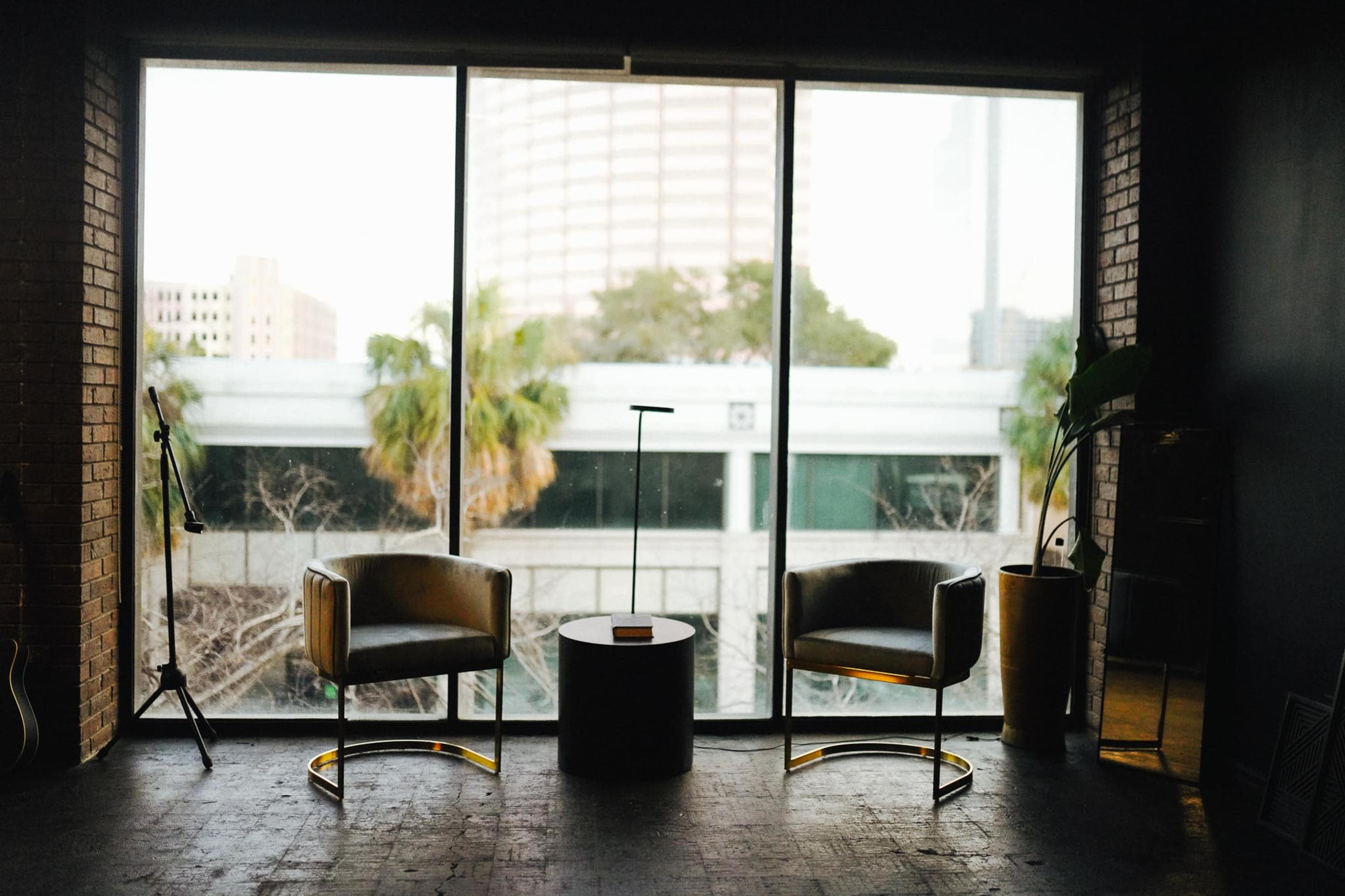 The image shows a minimalist interior with two chairs and a small round table, framed by large windows overlooking a cityscape with palm trees outside.