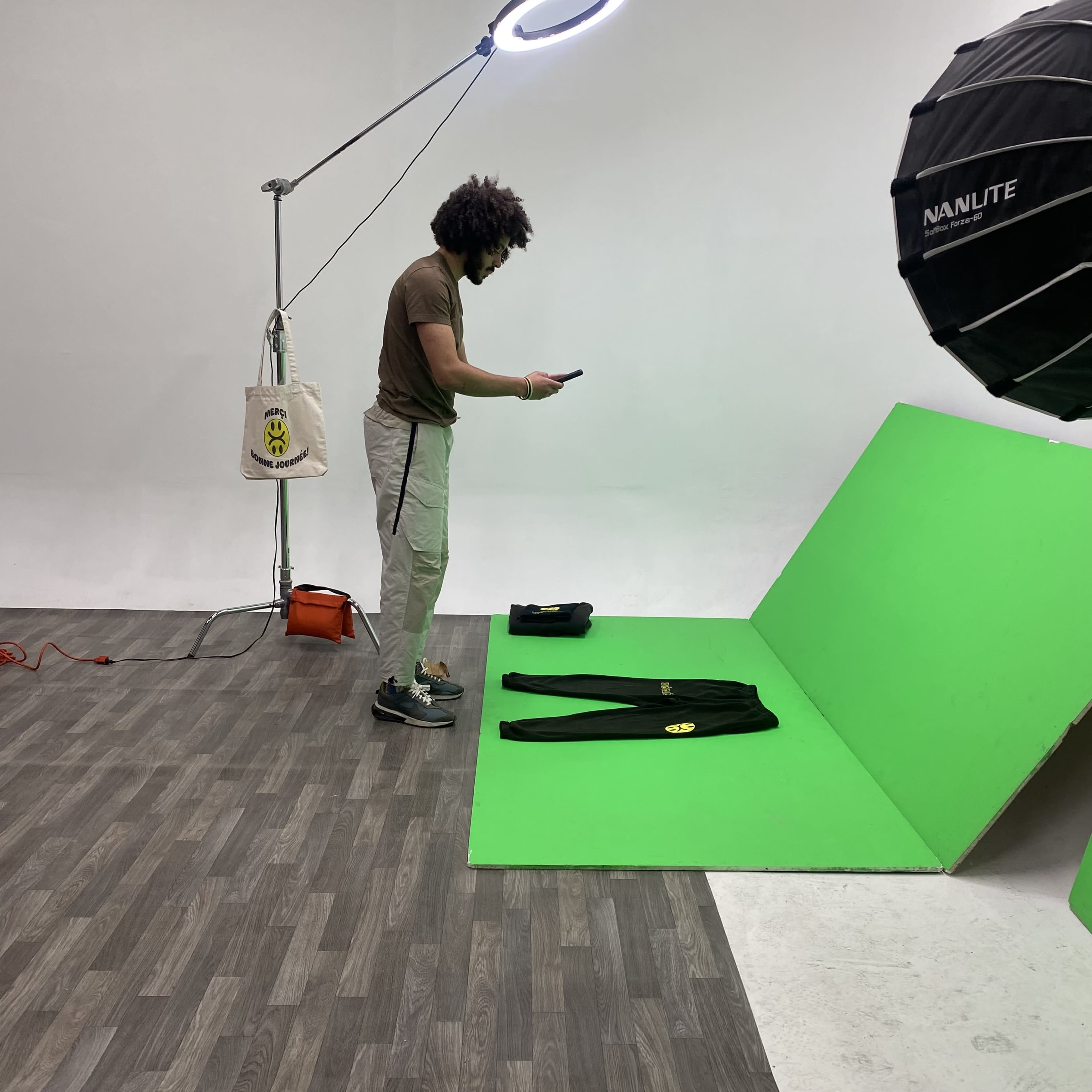 A person stands in a studio that features a green backdrop, while checking a phone next to laid-out clothing items.