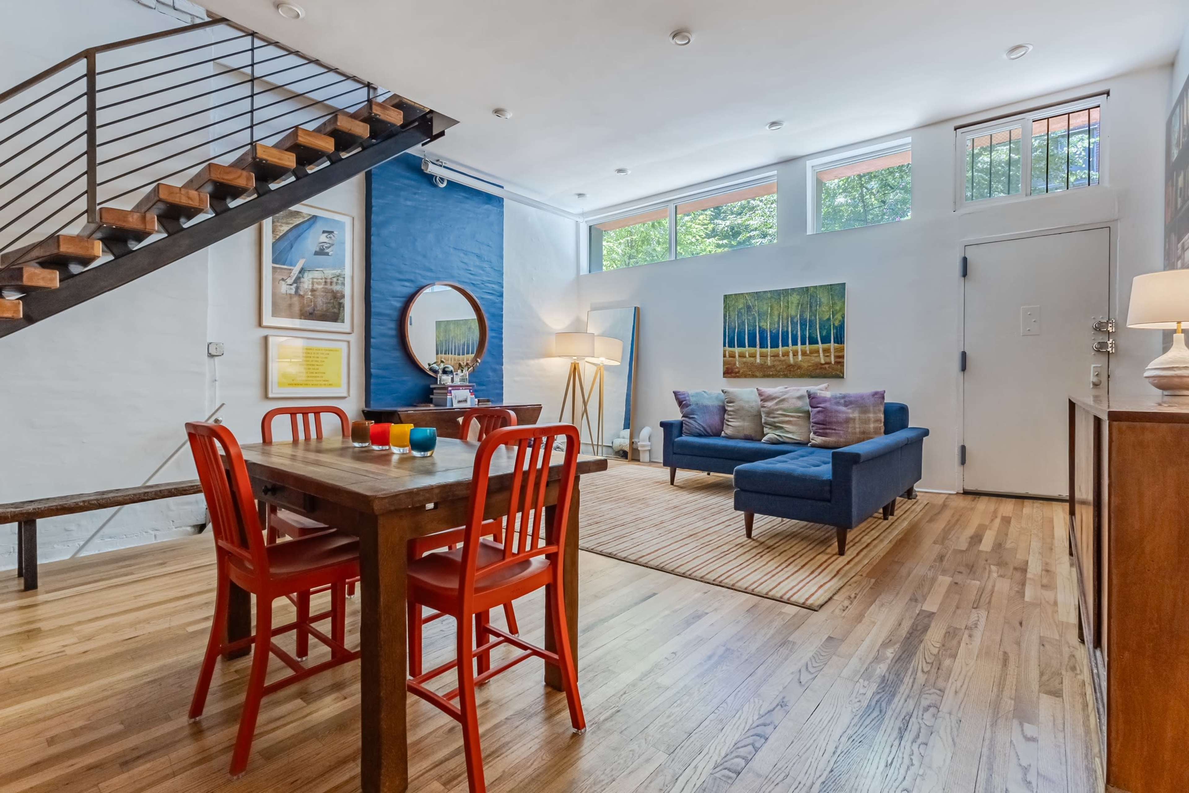 A modern living space with a dining area featuring a wooden table and red chairs, a blue sofa, and a staircase against a white wall.