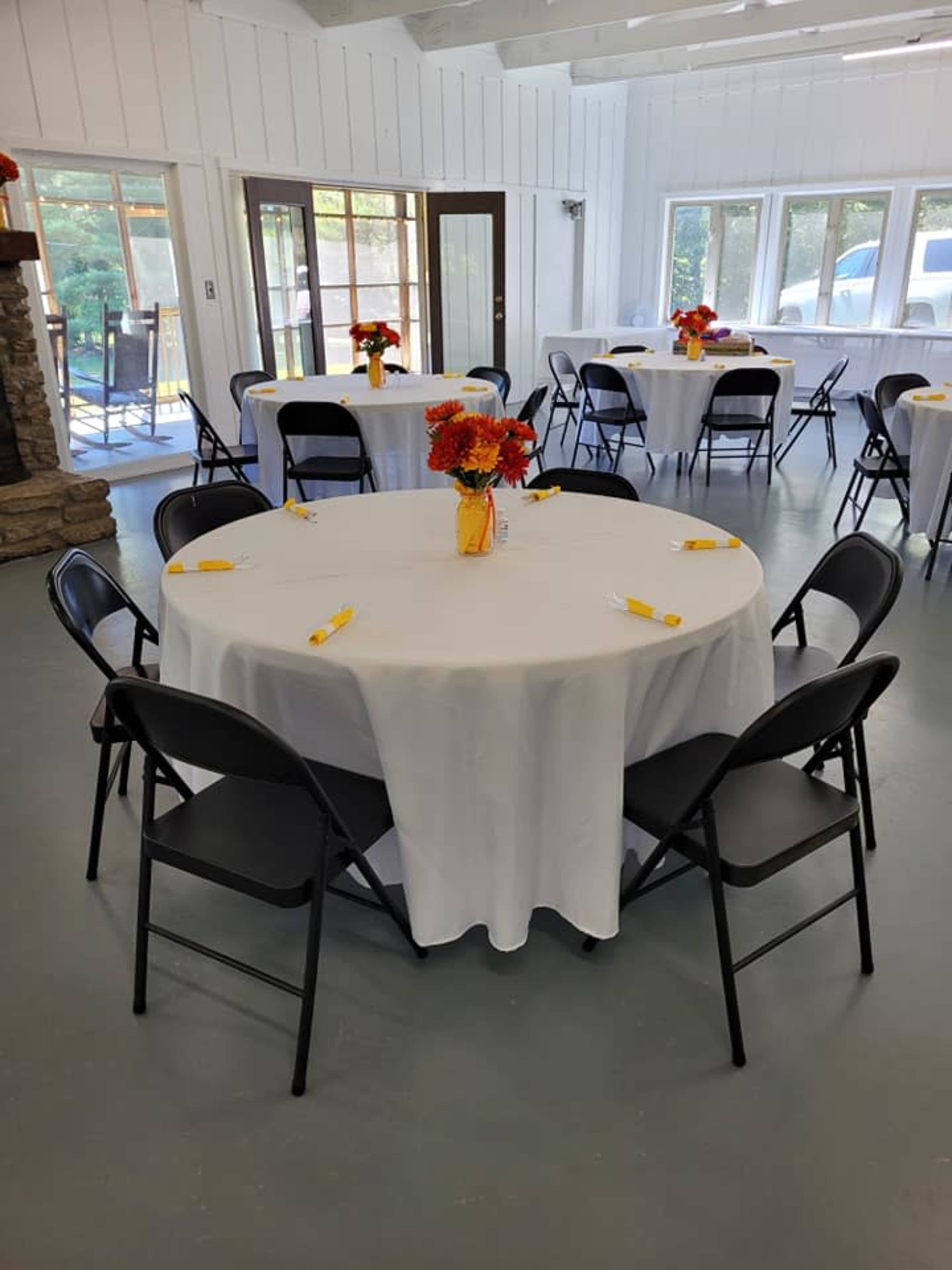 A set of round tables covered with white tablecloths is arranged in a bright room, each adorned with a small flower centerpiece and yellow utensils.