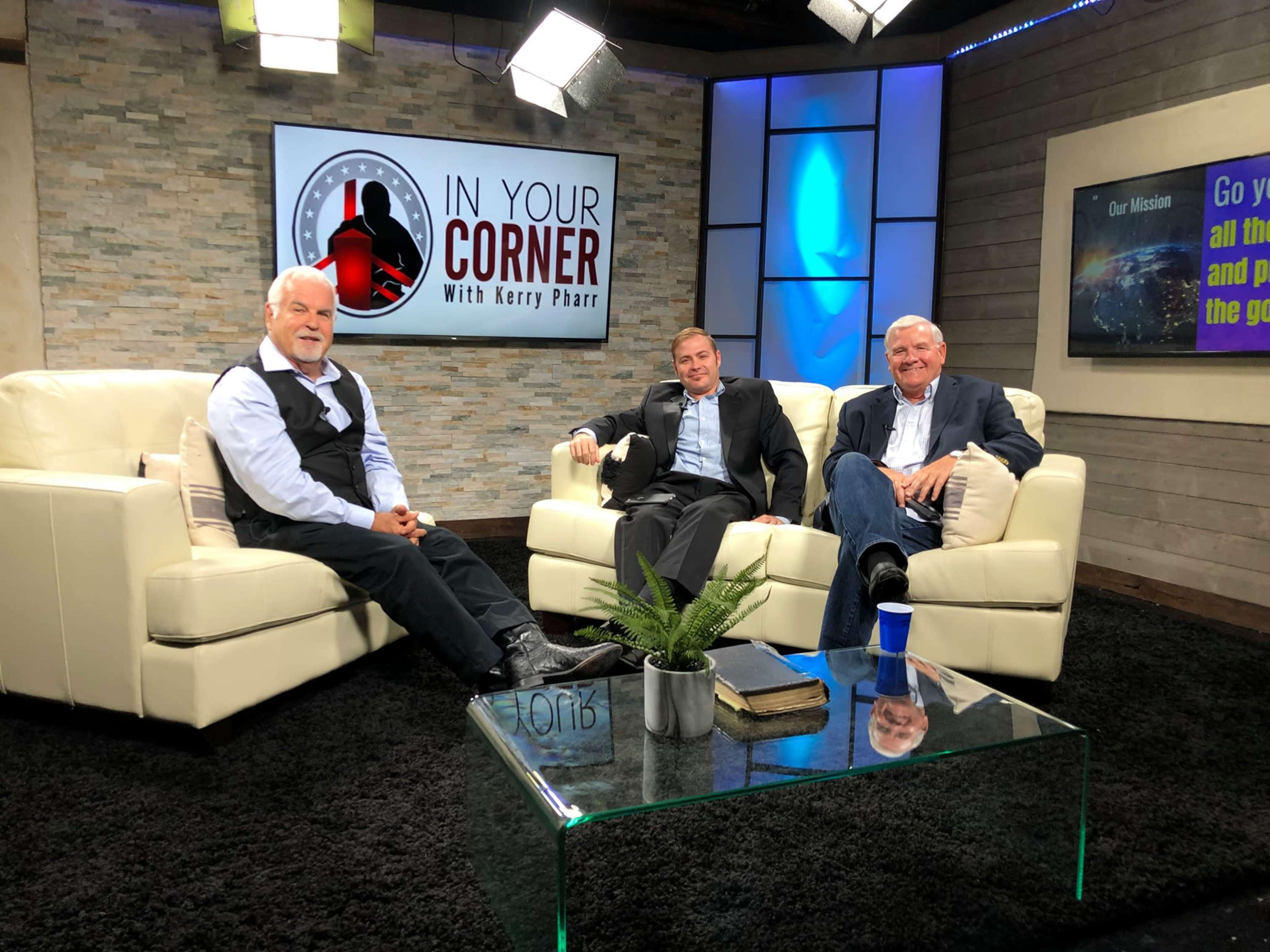 Three men are seated on a white couch in a well-lit studio, with a stone wall backdrop and a glass coffee table in front of them.