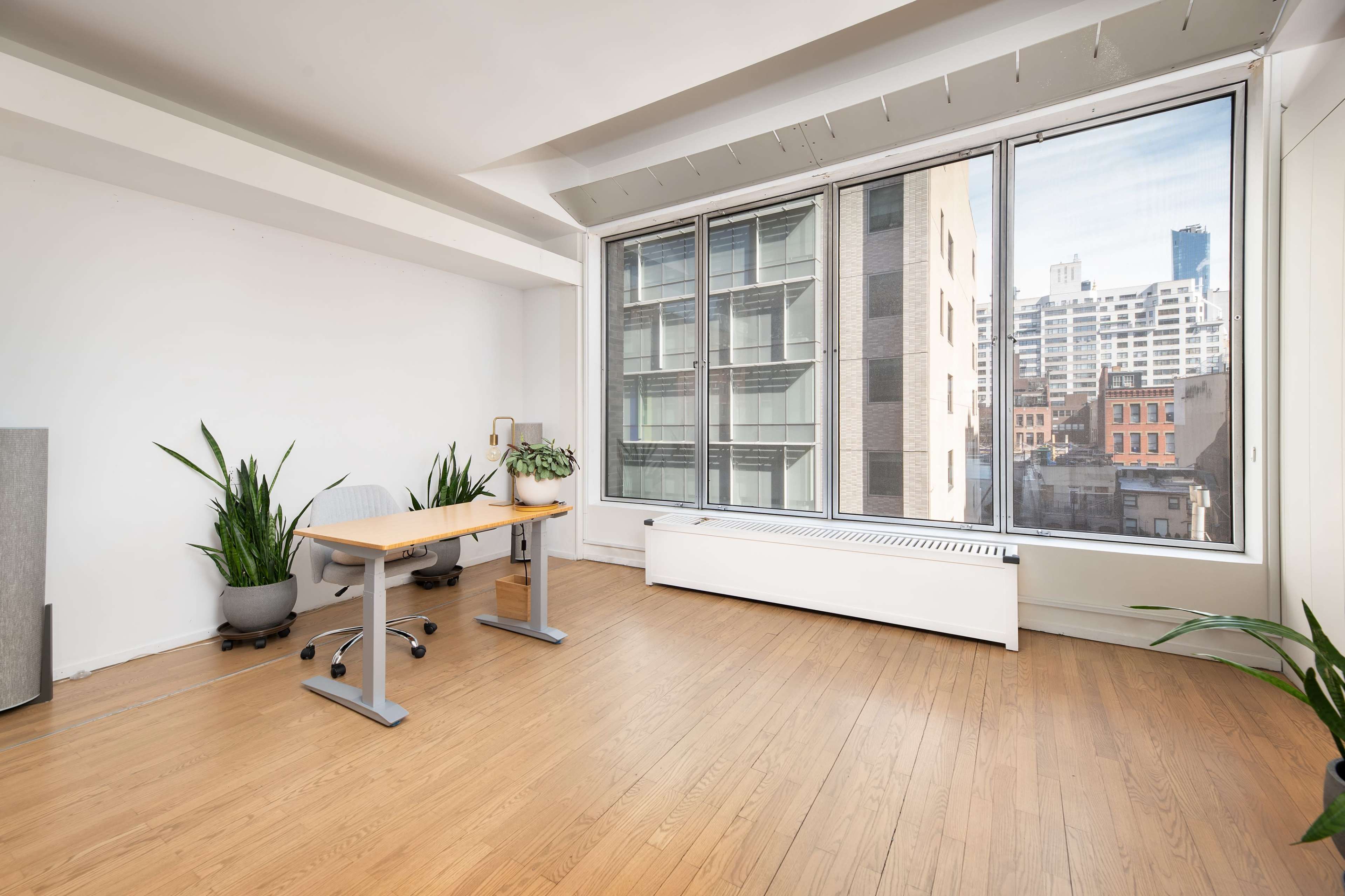An empty room with wooden flooring features a desk beside large windows overlooking a cityscape, with potted plants in the corners.