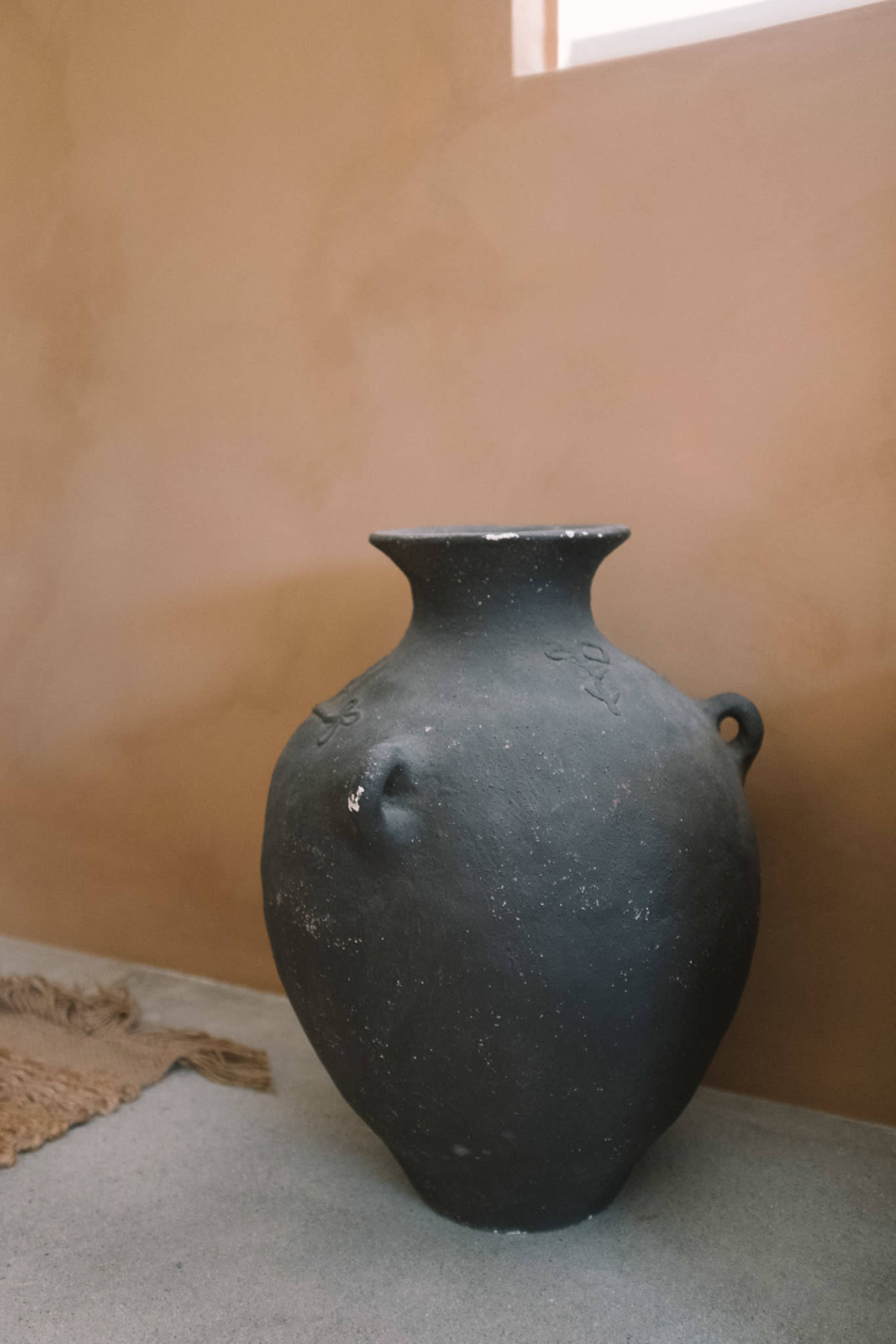 A dark pottery vase with a rounded body and a small handle stands against a clay-colored wall in a minimal interior setting.
