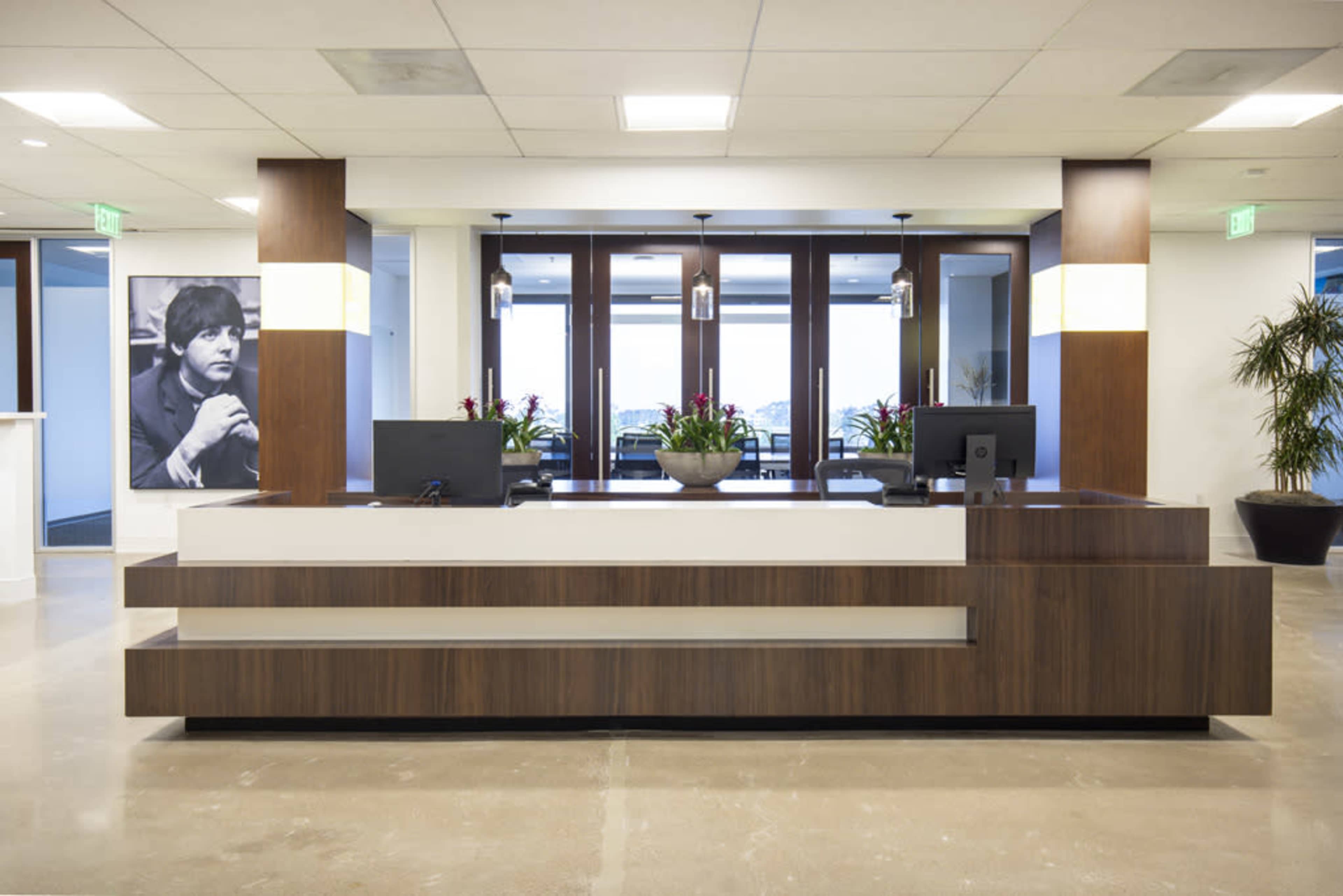 A modern reception desk in an office environment, featuring a large potted plant and a black-and-white photograph of a man on the wall.