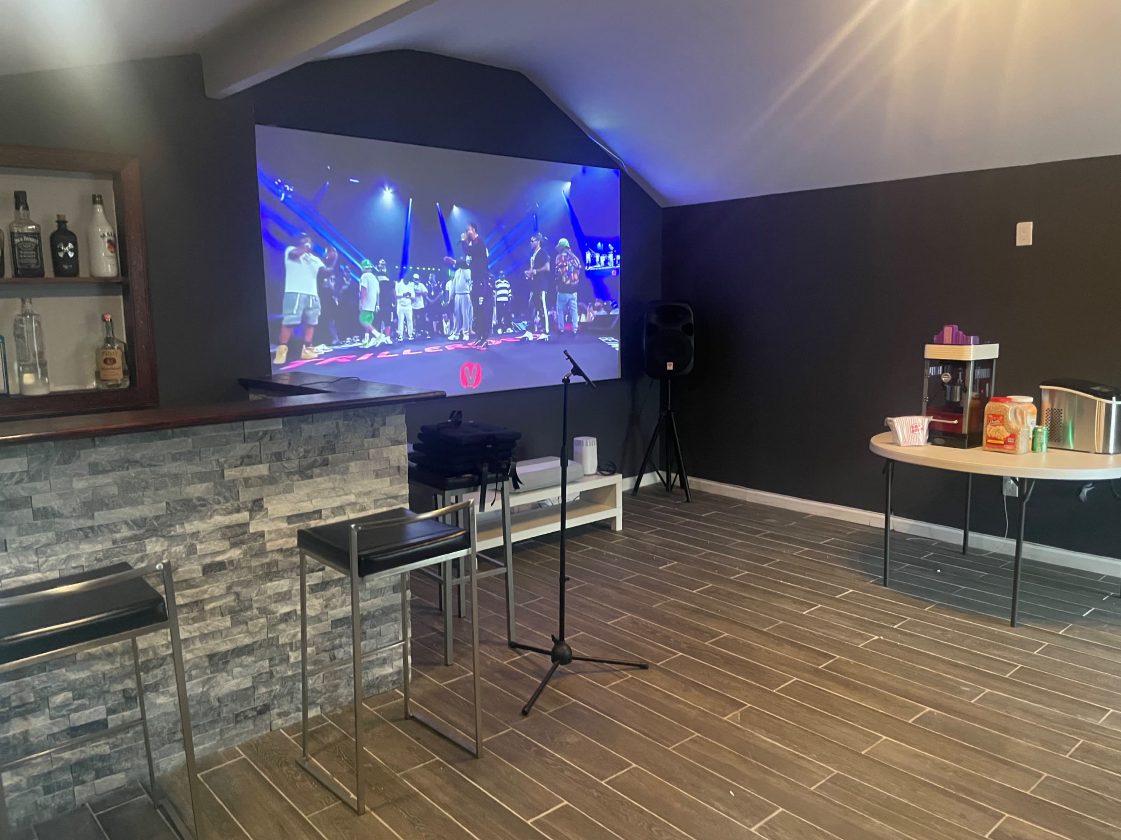 A bar area with a large screen displaying an event is shown, alongside a table with snacks and a microphone set up.