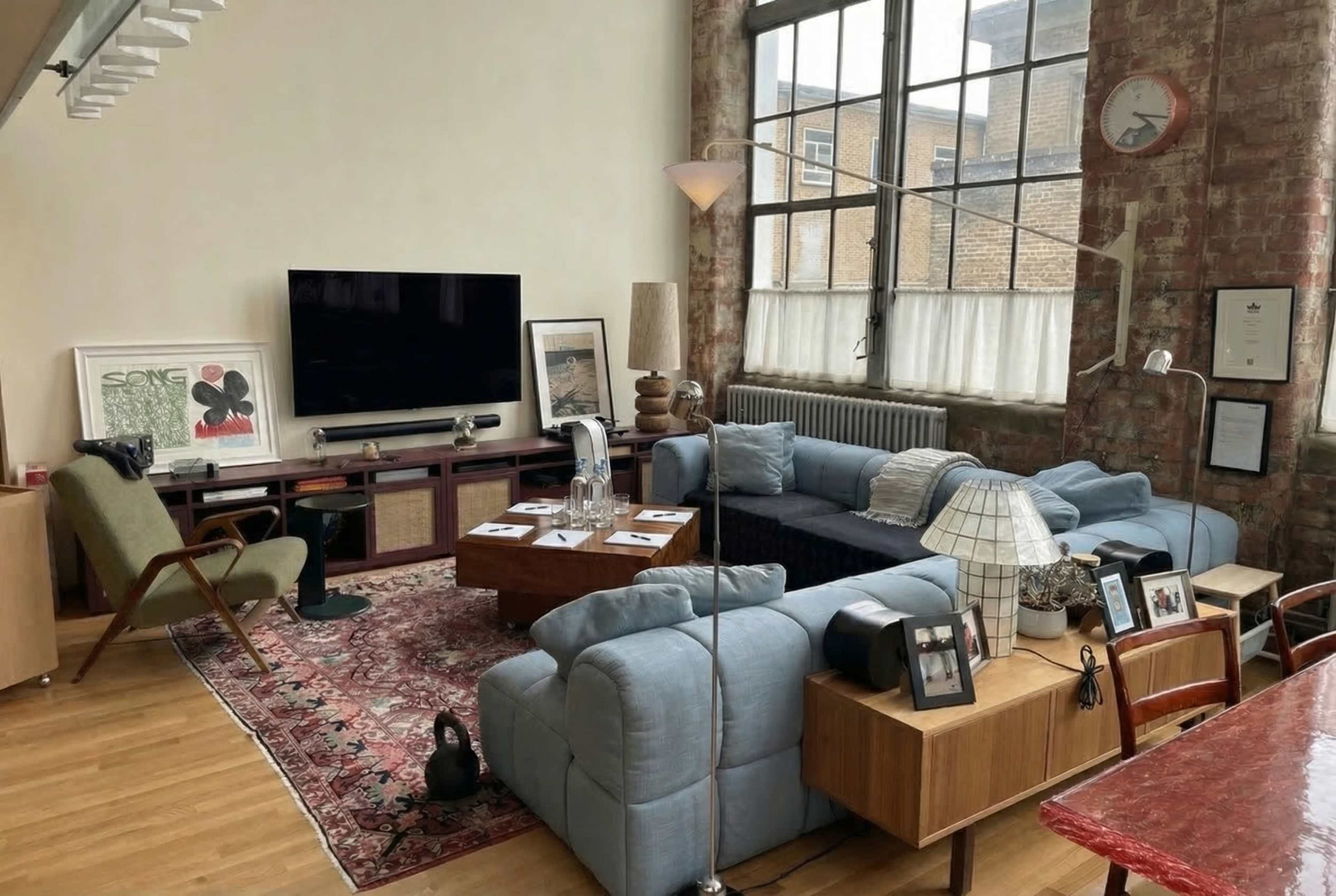 The image shows a modern living room with a large sectional sofa, a wall-mounted television, a wooden coffee table, and large windows letting in natural light, with exposed brick walls and various decorative items.