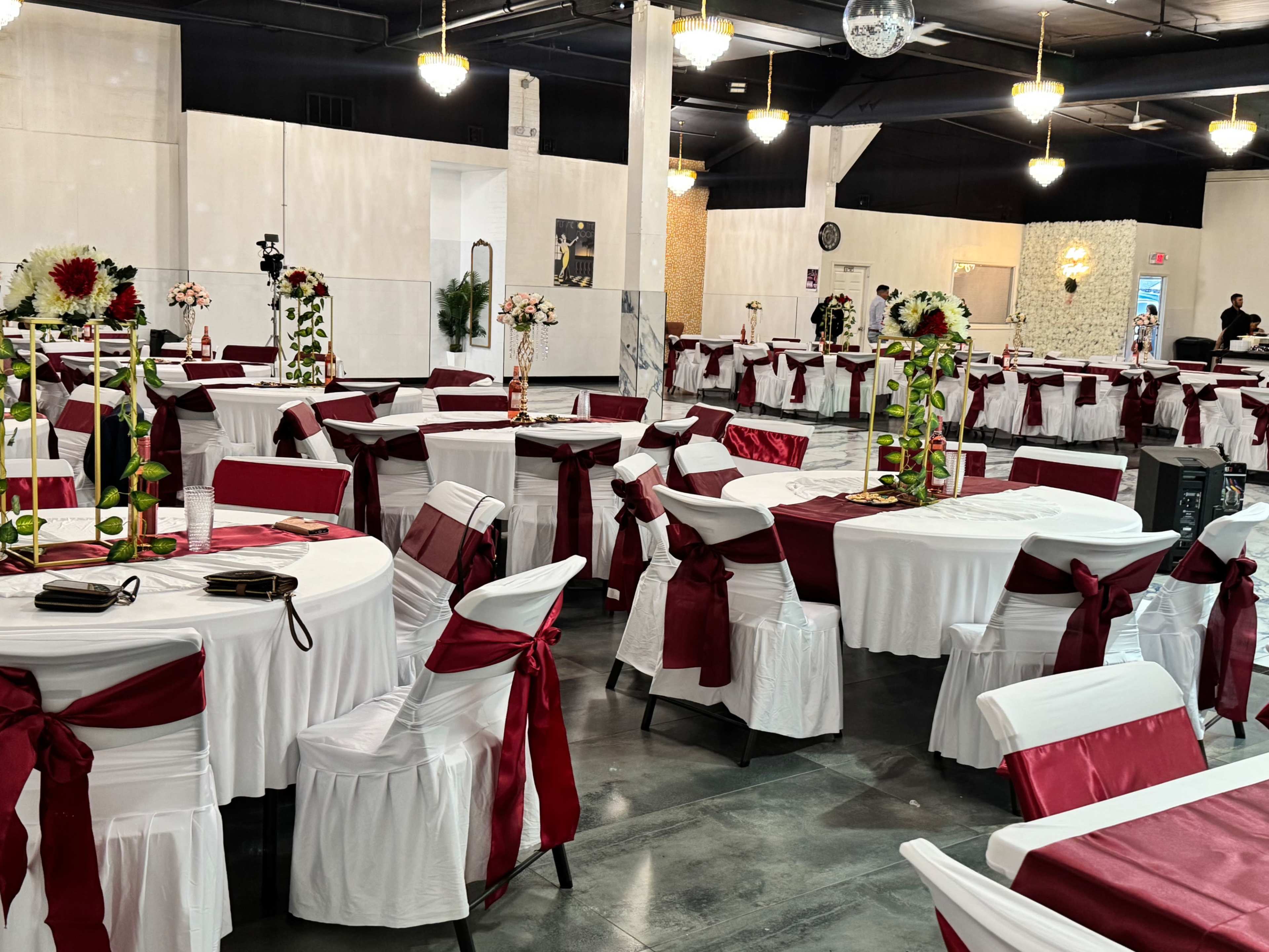 The image shows a spacious banquet hall set for an event, featuring round tables with white tablecloths and burgundy accents, surrounded by decorated chairs.